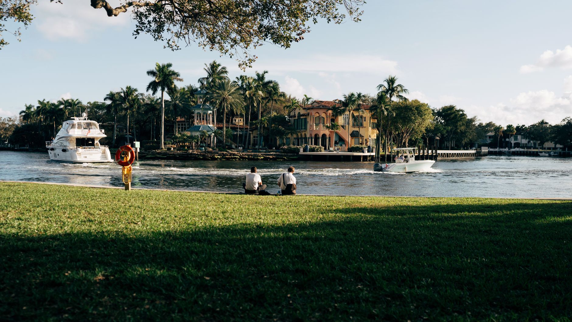A peaceful riverside view featuring palm trees, boats, and people relaxing by the water.