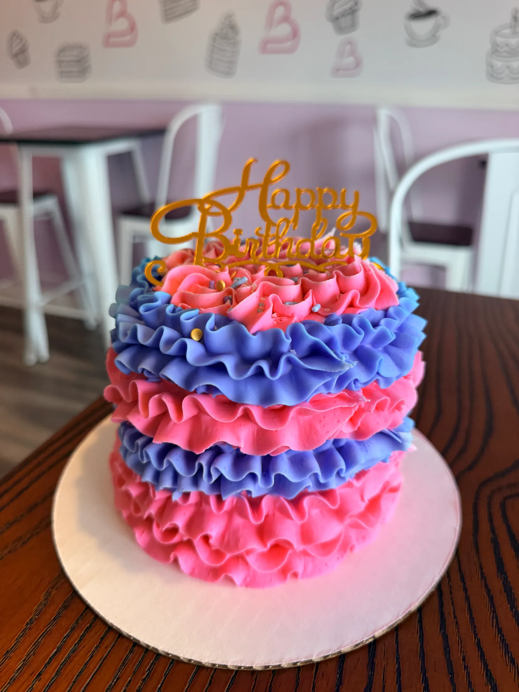 Close-up of a ruffled pink and blue buttercream birthday cake with a gold Happy Birthday topper at Brenda's Cake Shop