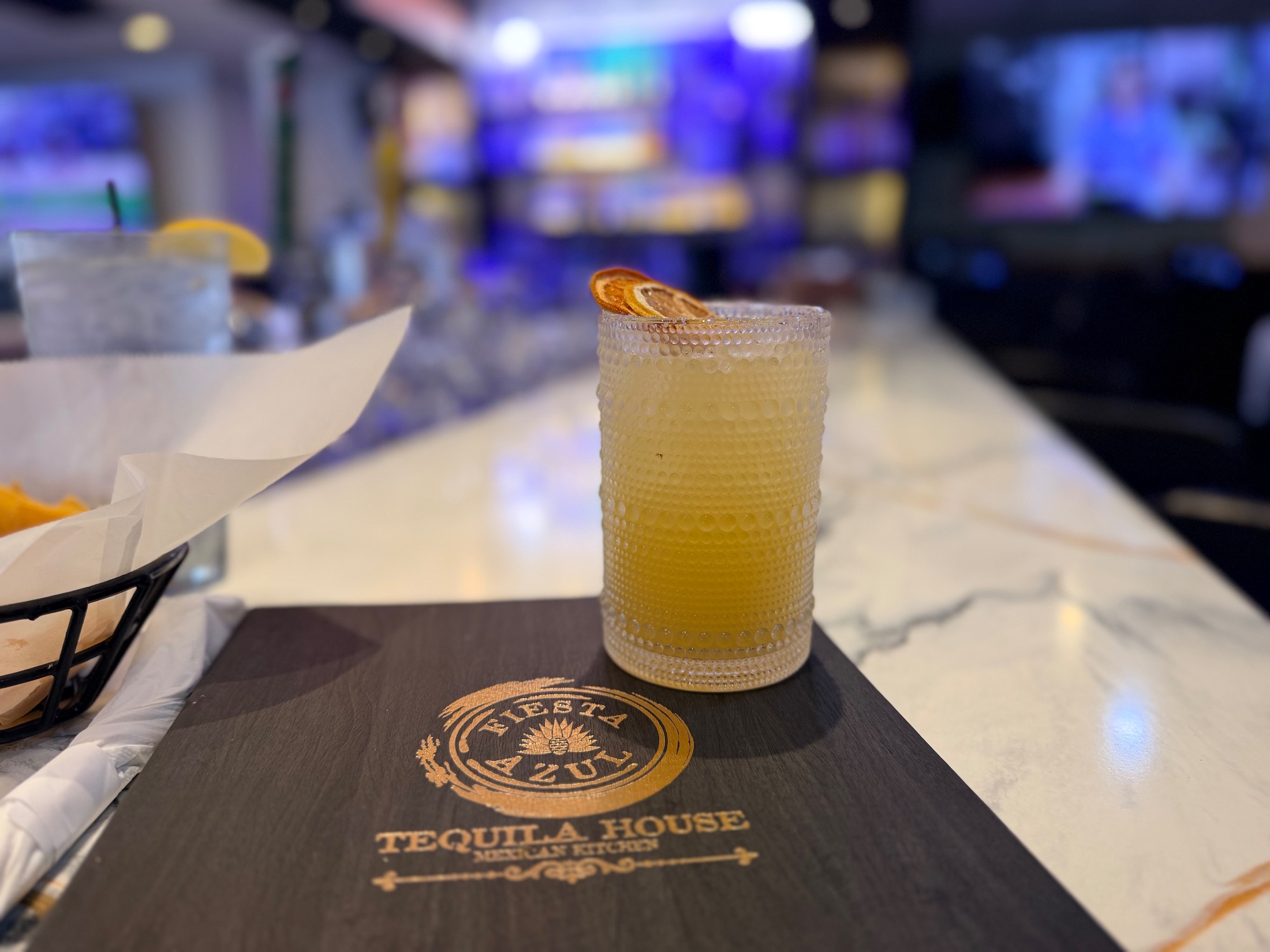 House margarita in a textured glass with dried orange garnish next to a Fiesta Azul Tequila House menu on the bar