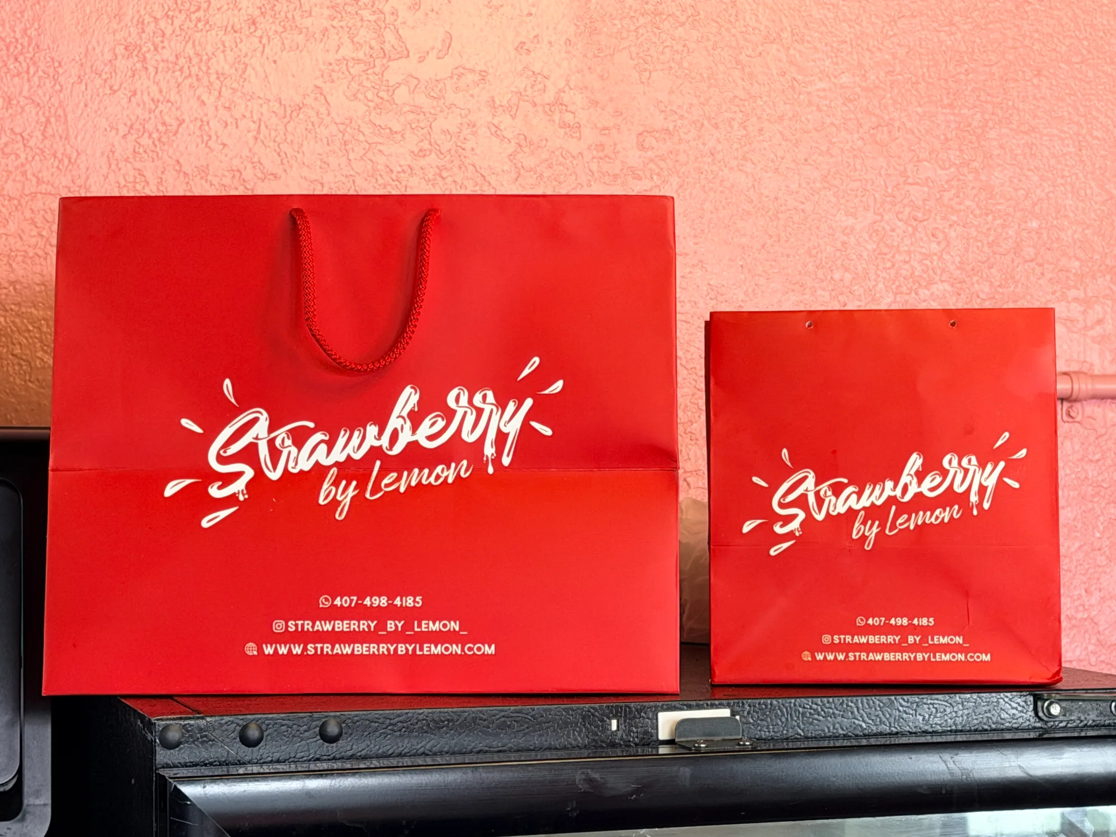 A slice of plain cheesecake on a wooden board in front of a Strawberry by Lemon striped takeout bag