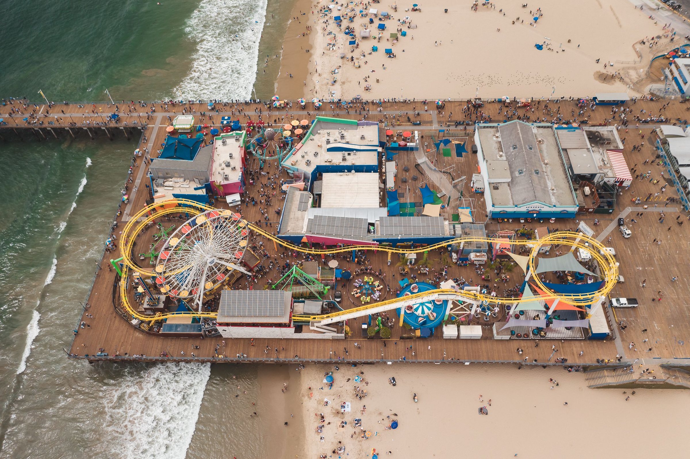 Aerial view looking straight down at a pier over the ocean, with a Ferris wheel, a roller coaster track and other rides on the deck and a crowded sandy beach