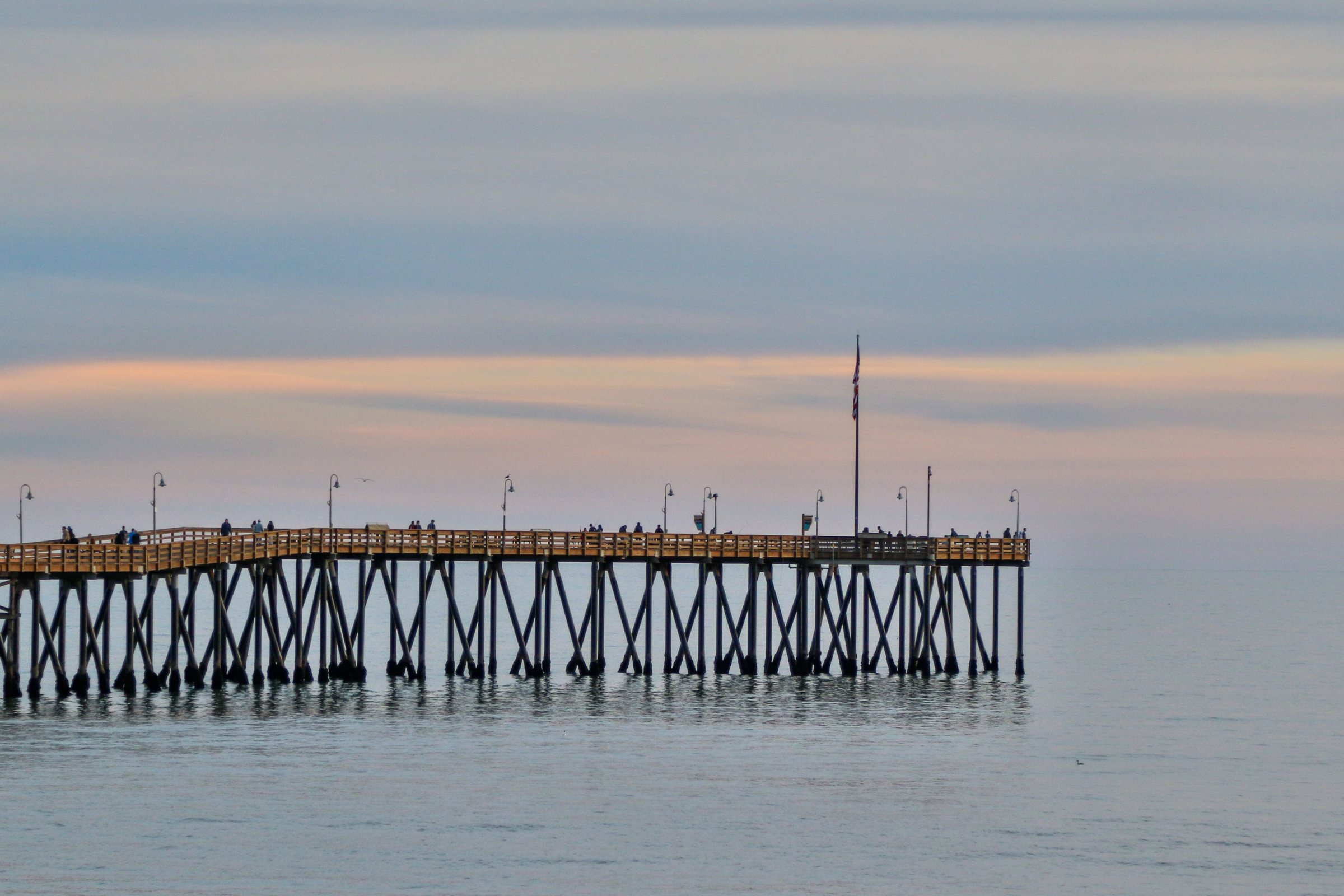 A long wooden pier on many thin pilings across calm gray-blue water at dusk, with pink and orange clouds above the horizon