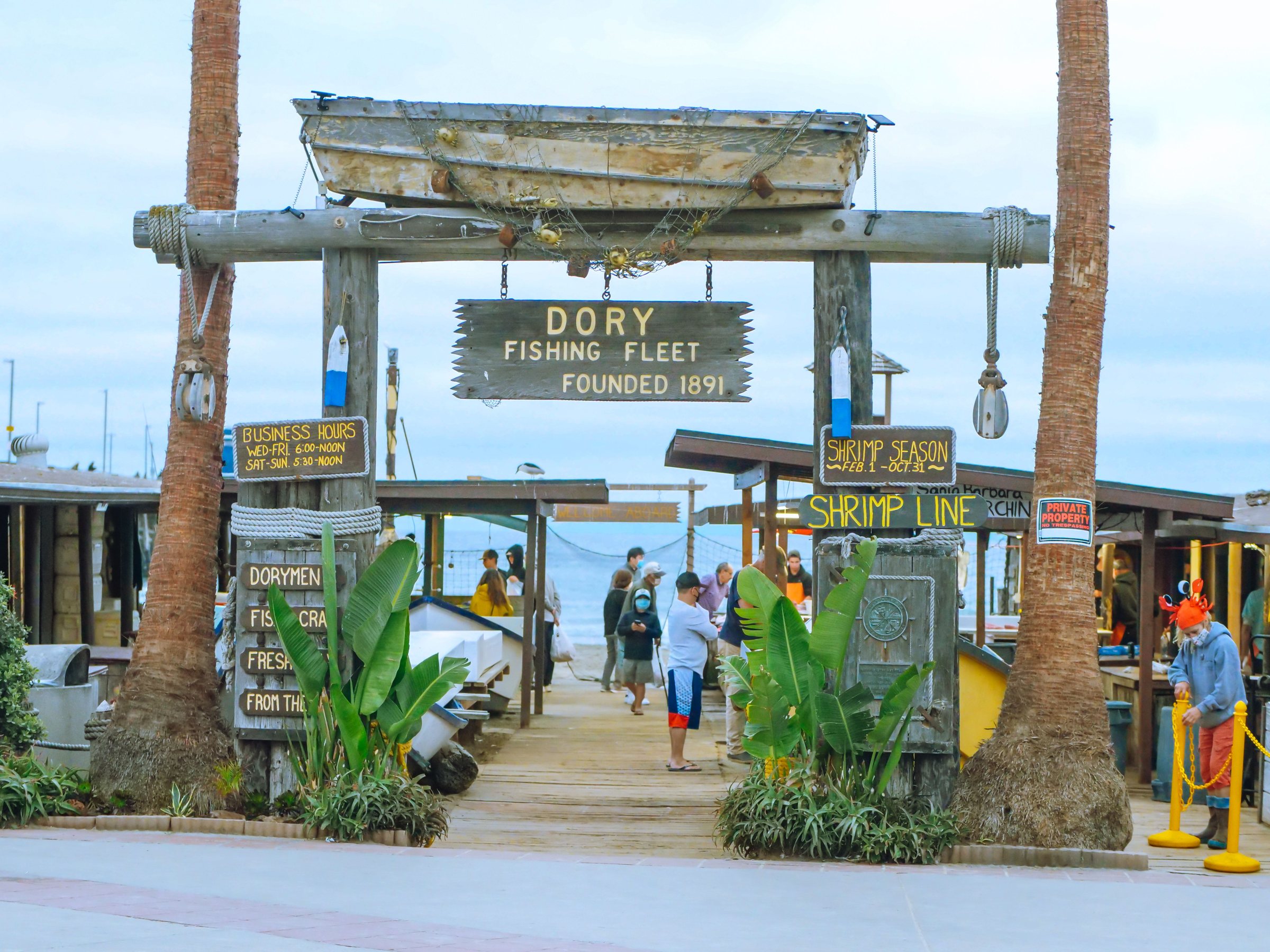 A wooden boat mounted on top of a Dory Fishing Fleet sign between two palm trees, with people walking below