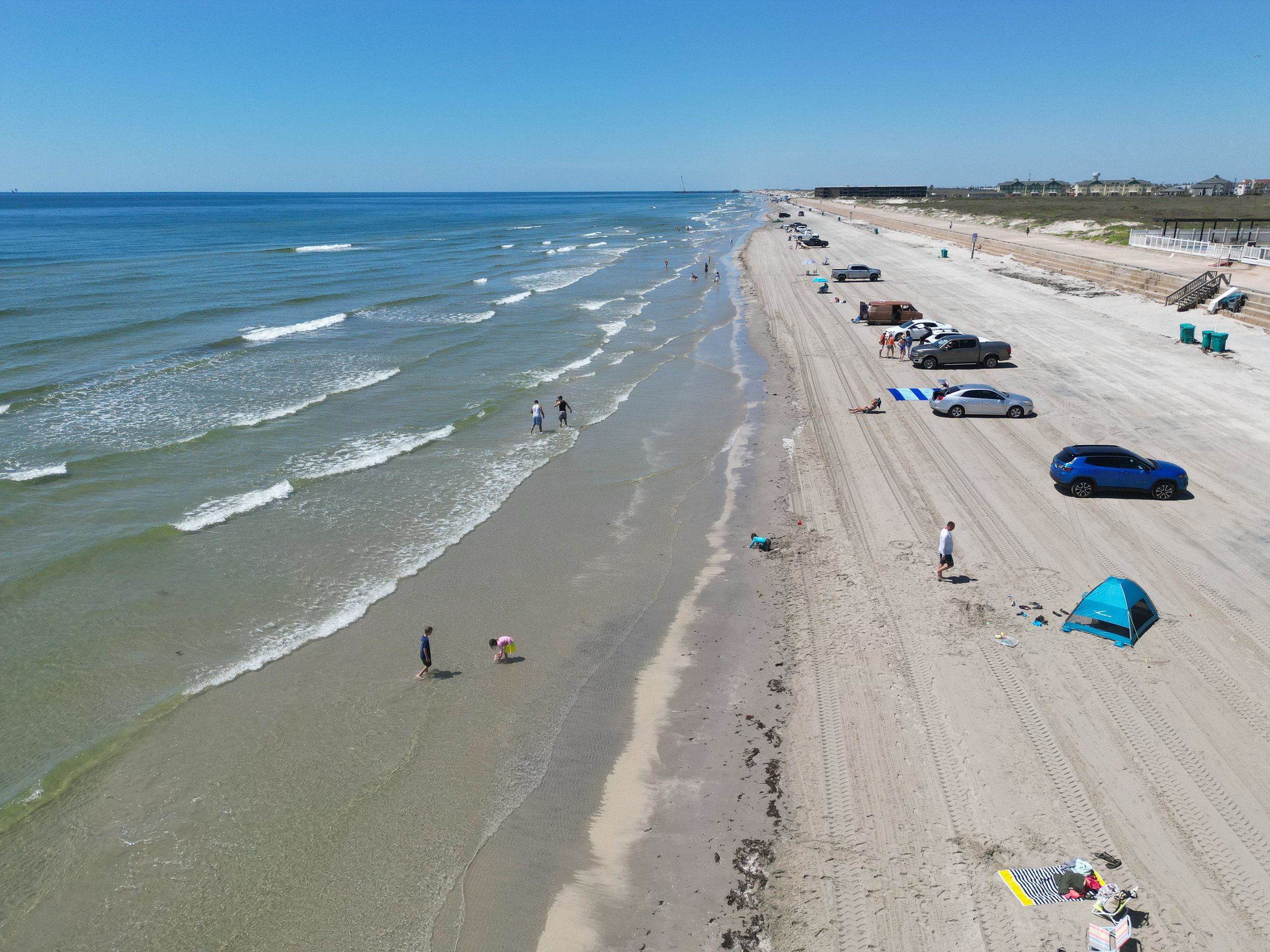 Aerial view along a long straight sandy beach with green-blue water and white surf on one side and cars, a blue tent and a few people on the wide sand