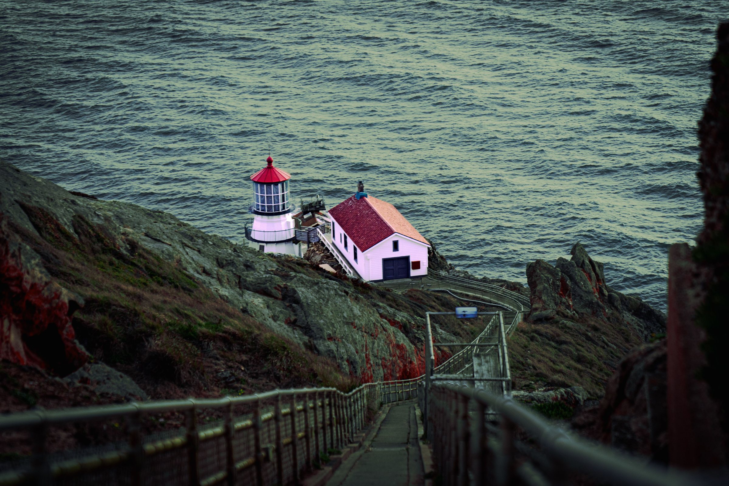 A small white lighthouse with a red top and a white building with a red roof sit on a rocky green point above dark green ocean water, with a long stairway with metal railings leading down the steep cliff toward them