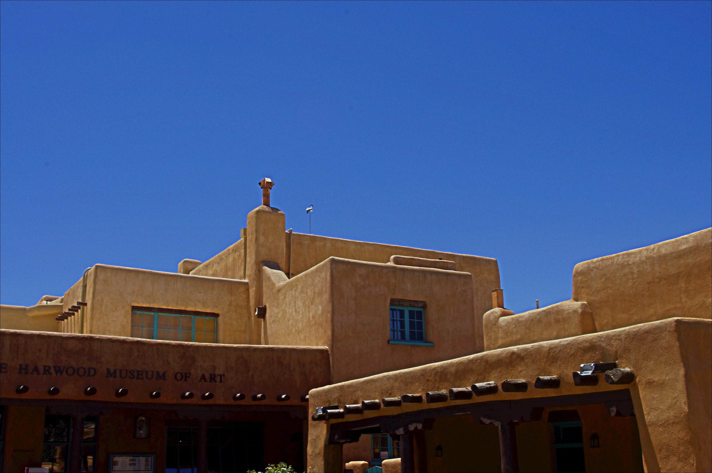 A tan adobe building with wooden vigas and a small tower under a deep blue sky, the Harwood Museum of Art on Ledoux Street