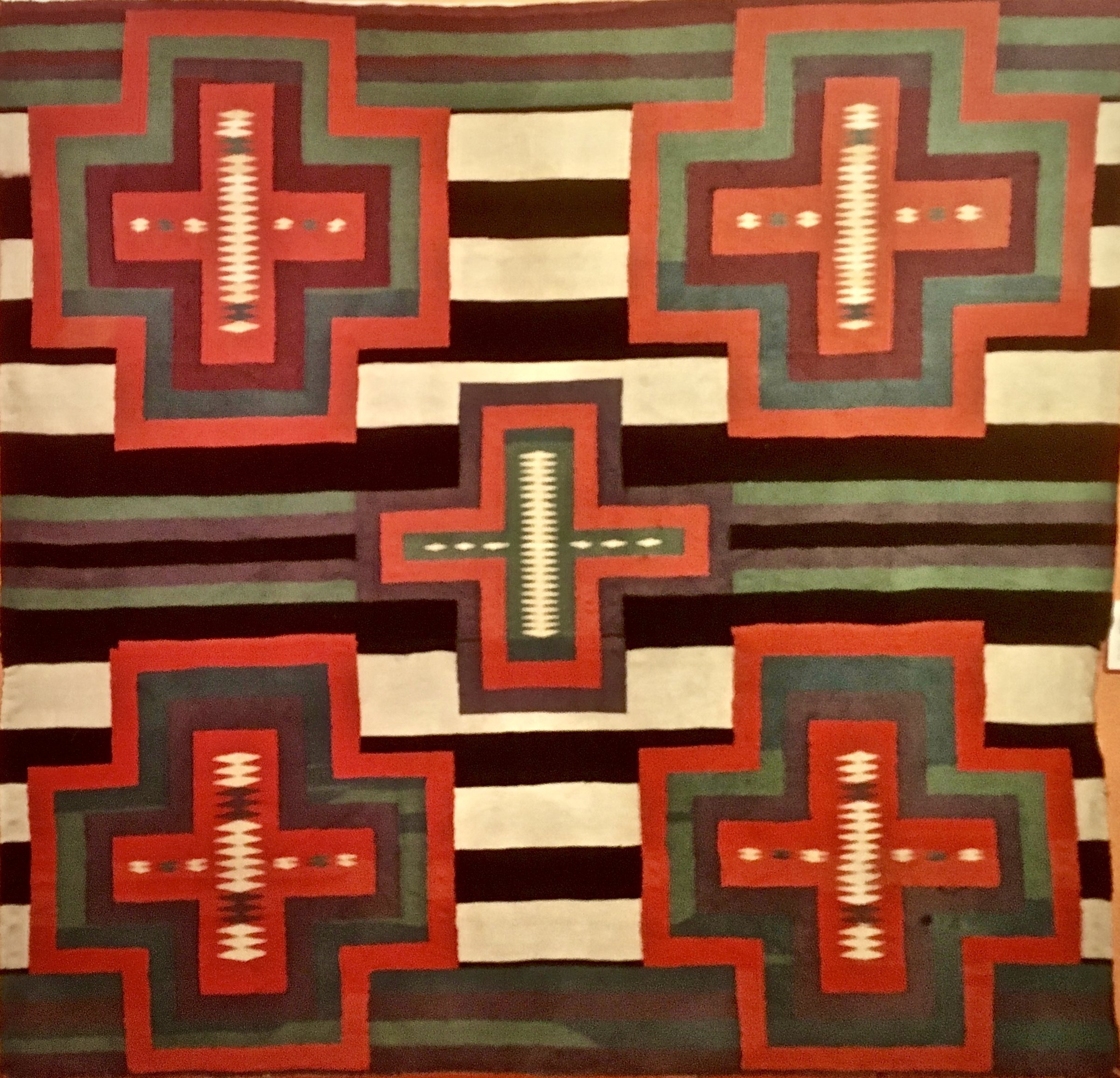 A woven Navajo blanket with red stepped crosses on bands of black, white and red, shown at the Millicent Rogers Museum