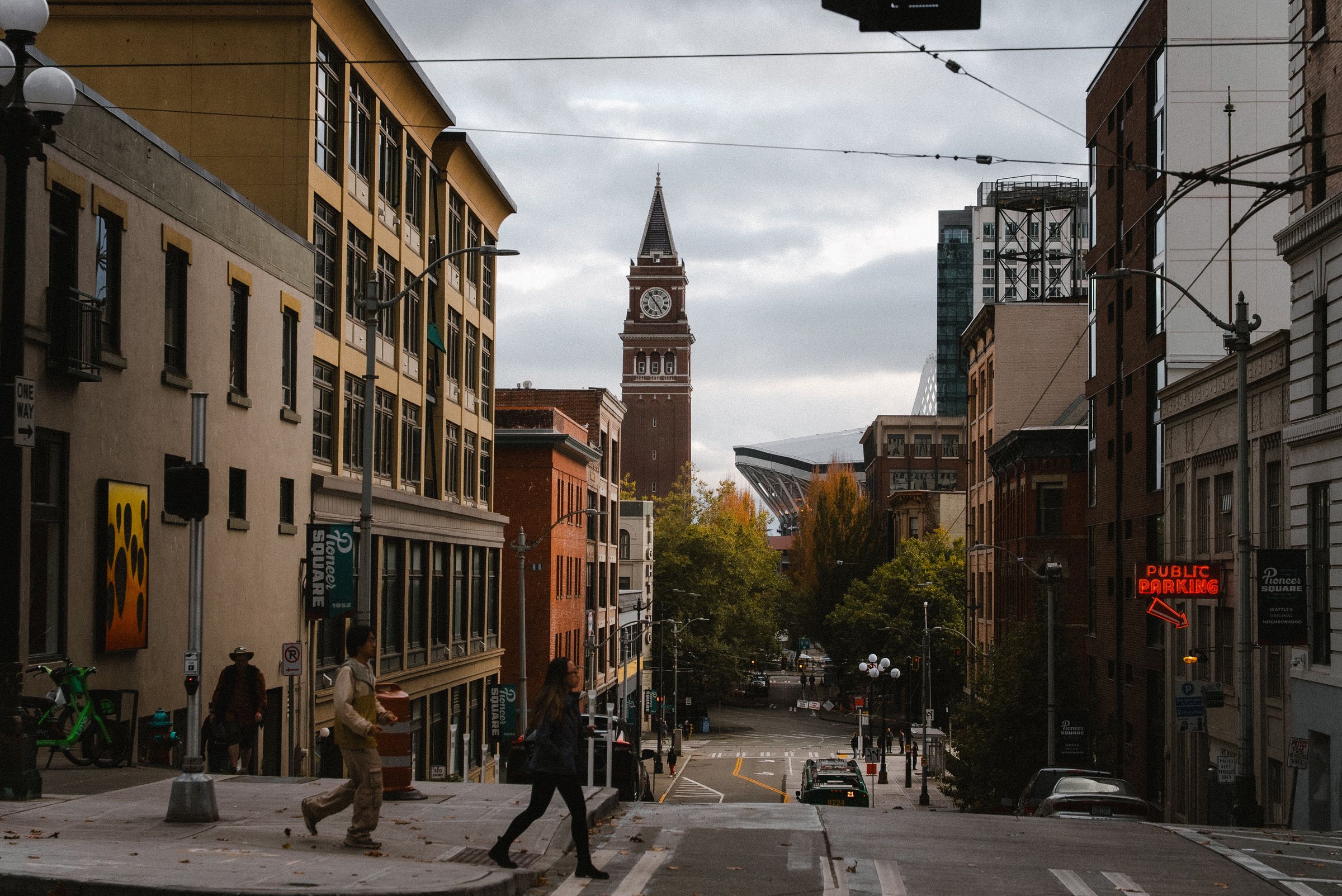 Two pedestrians crossing a street lined with brick and stone buildings, with green banners reading Pioneer Square and a tall brick clock tower with a pointed roof at the far end