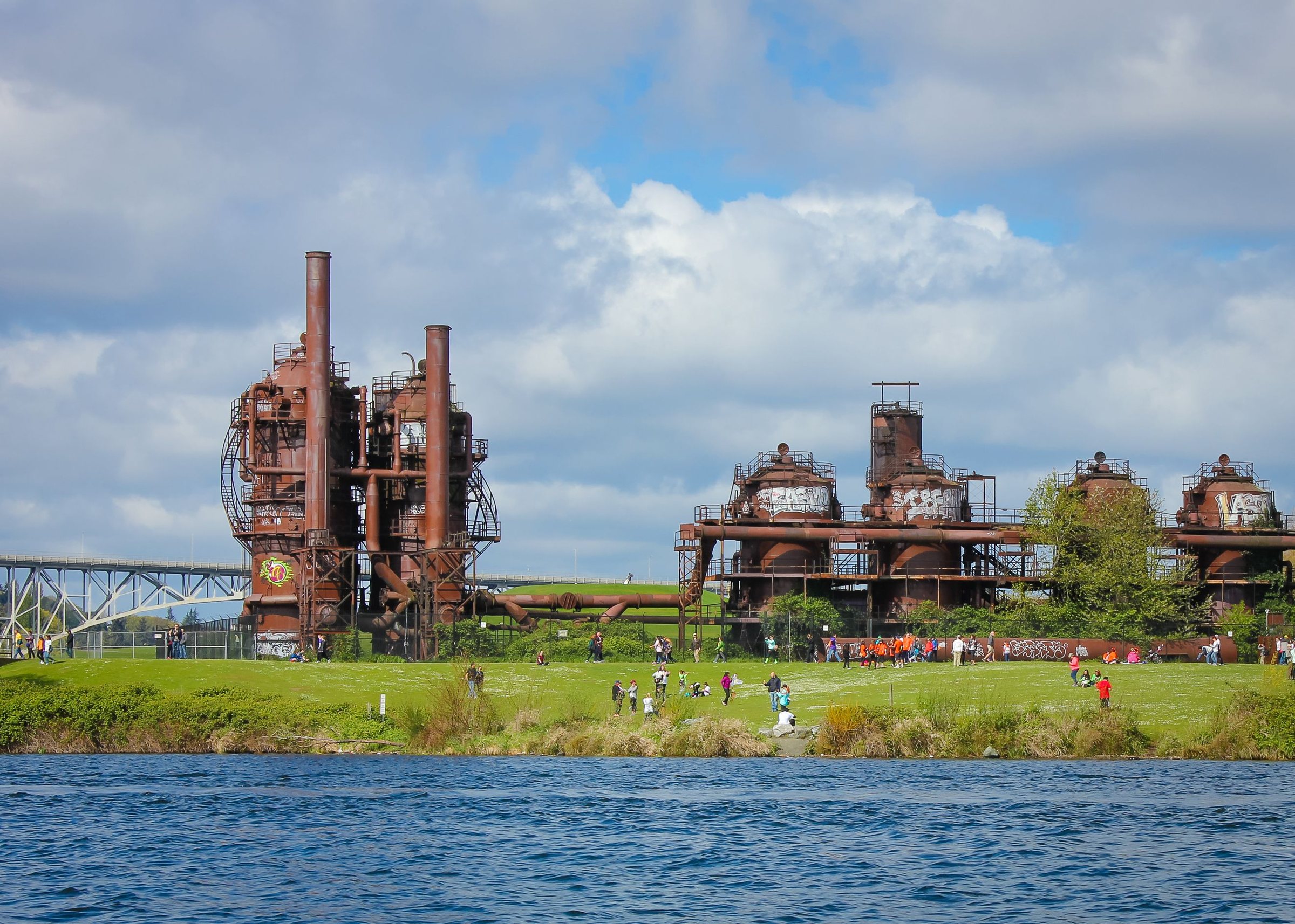 Tall rust-colored industrial towers and pipes beyond a strip of green lawn with people on it, seen across water