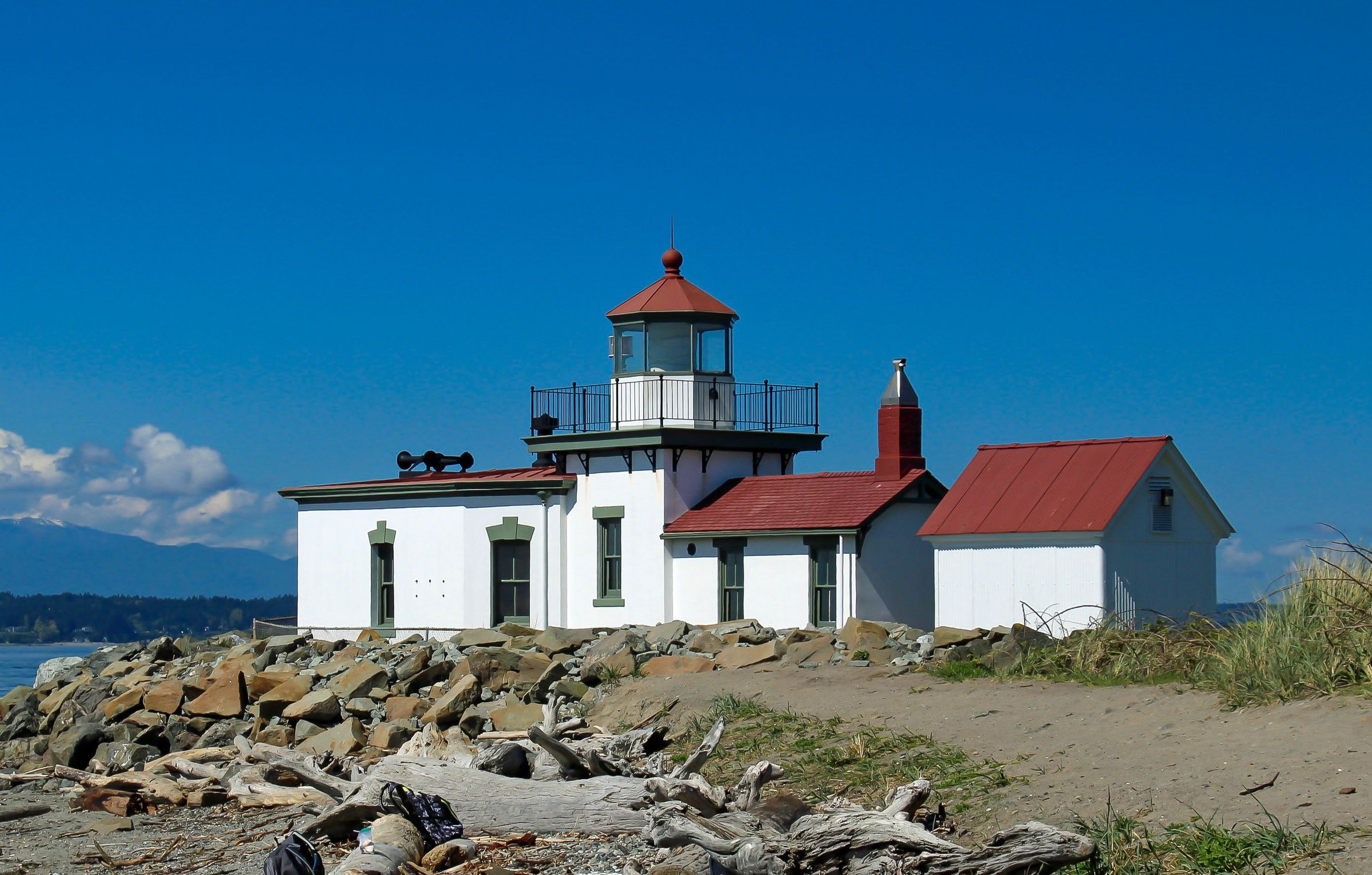 A white lighthouse with a red lantern roof next to white buildings with red roofs, with rocks and driftwood in front and a clear blue sky