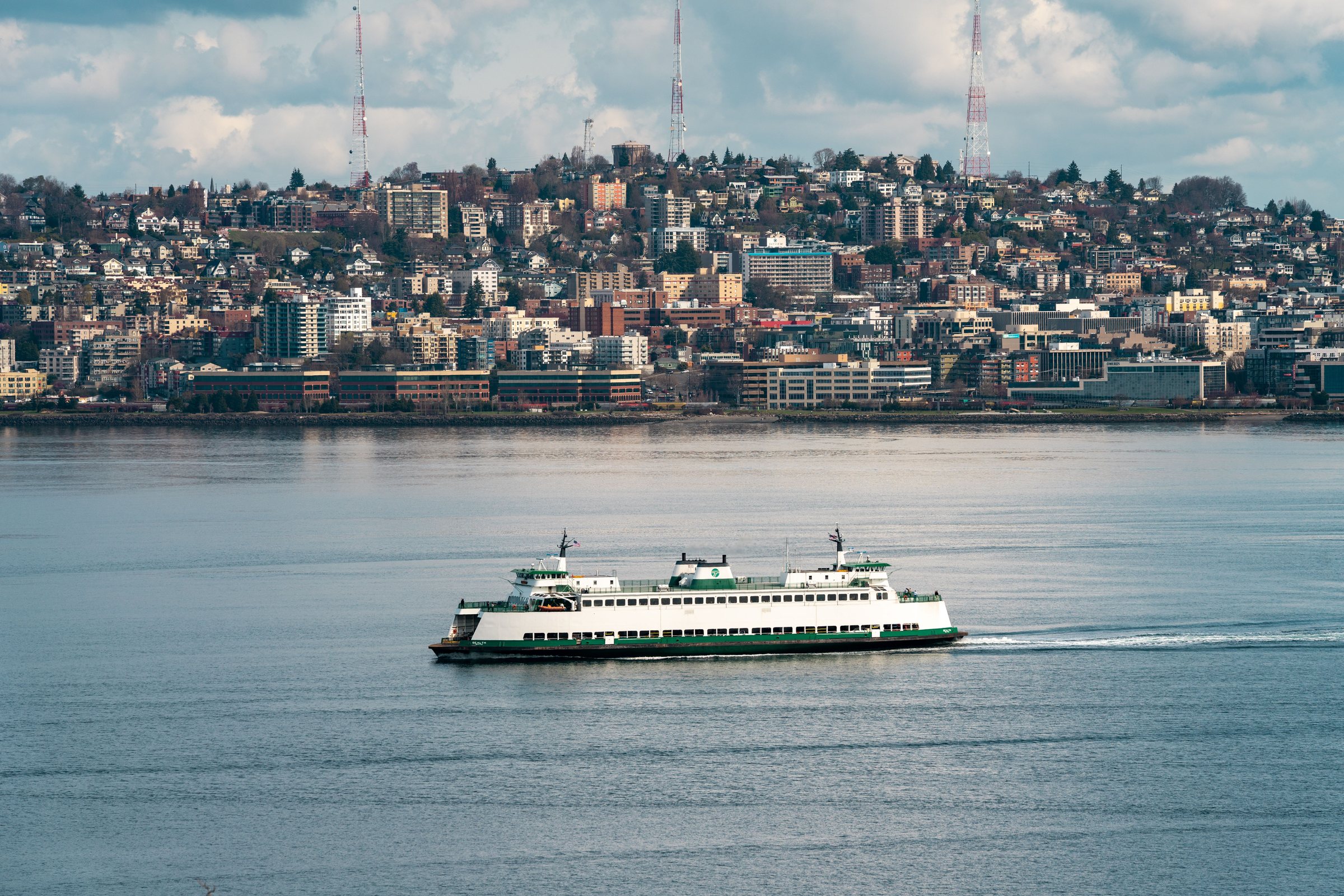 A green and white ferry on calm water, with a hillside covered with houses and buildings and radio towers on the hilltop behind it