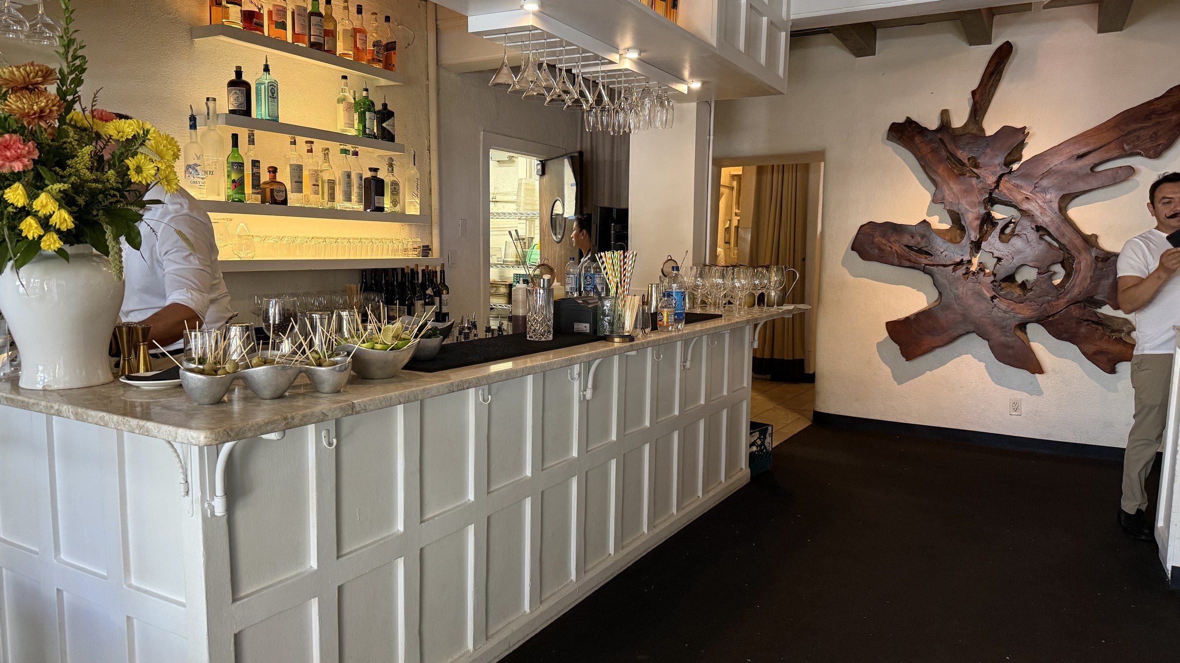 A white paneled bar with lit shelves of bottles, flowers in a white vase and a large carved wood sculpture on the white wall