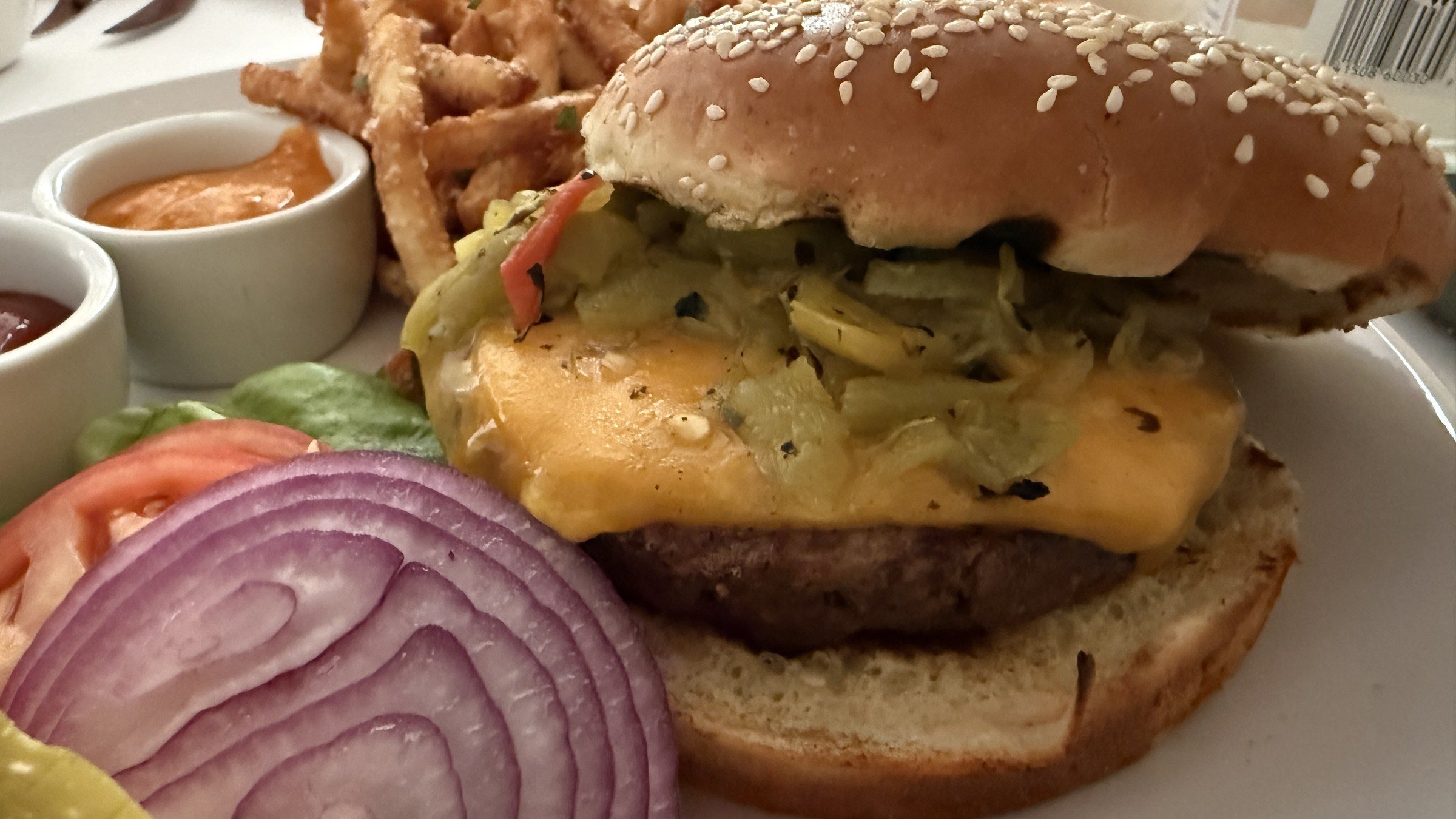 A cheeseburger with melted cheddar and chopped green chile on a sesame bun beside red onion, tomato and fries
