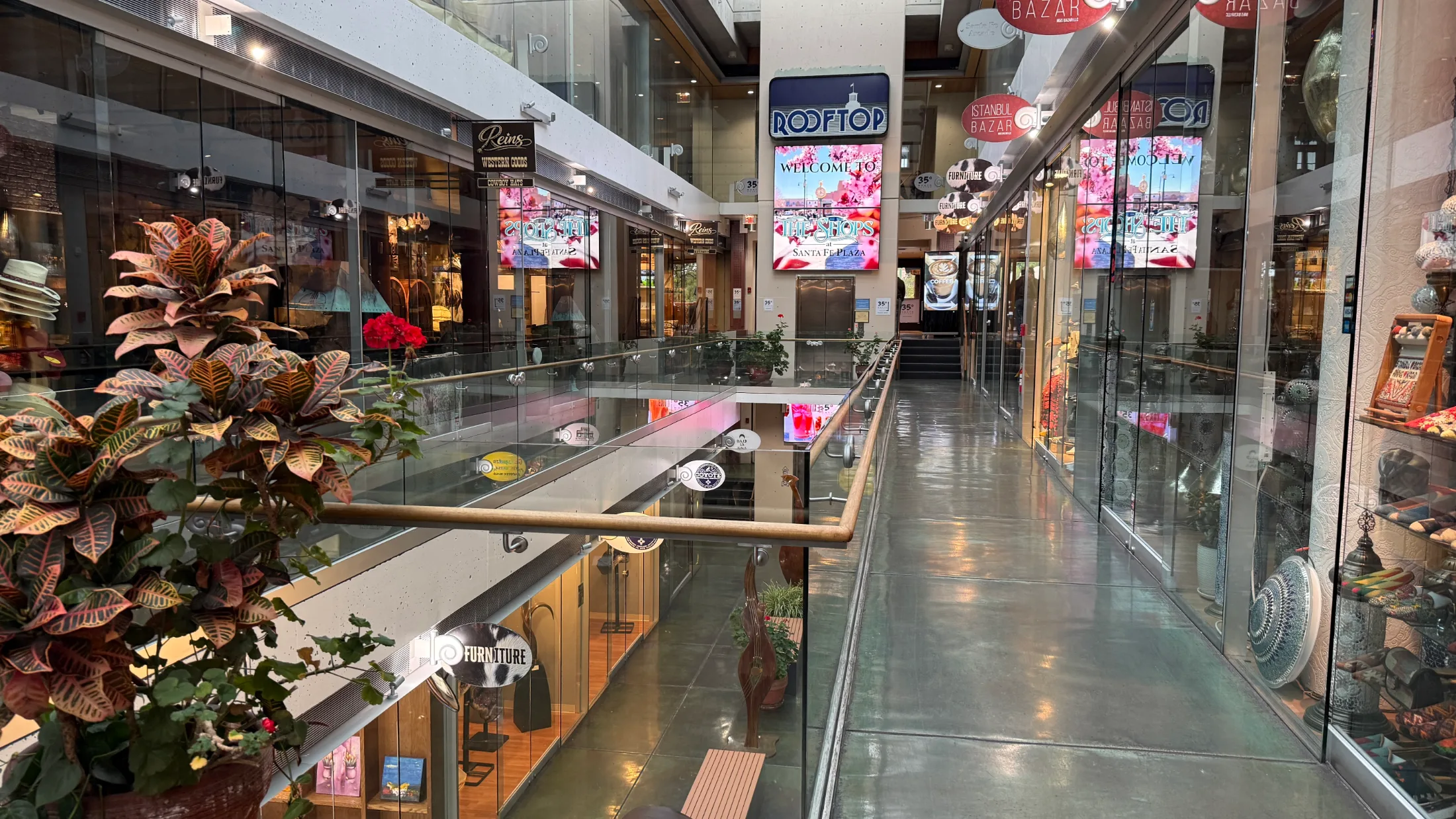 The upper level of the Shops at Santa Fe Plaza looking toward the Welcome screens and Rooftop sign, with Reins Western Goods and Istanbul Bazar visible on either side and potted plants along the glass railing