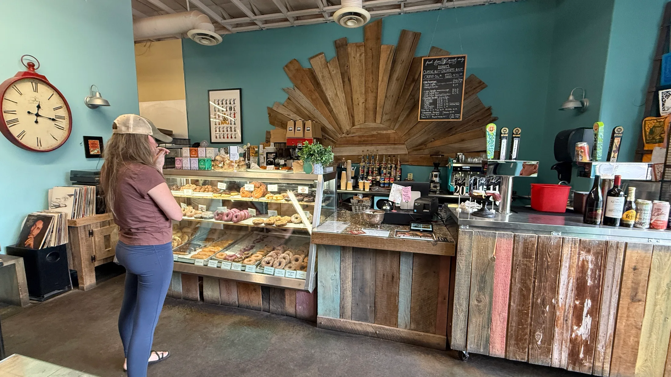 Inside Whoo's Donuts, a customer considers the glass display case filled with donuts beneath a reclaimed wood sunburst wall feature