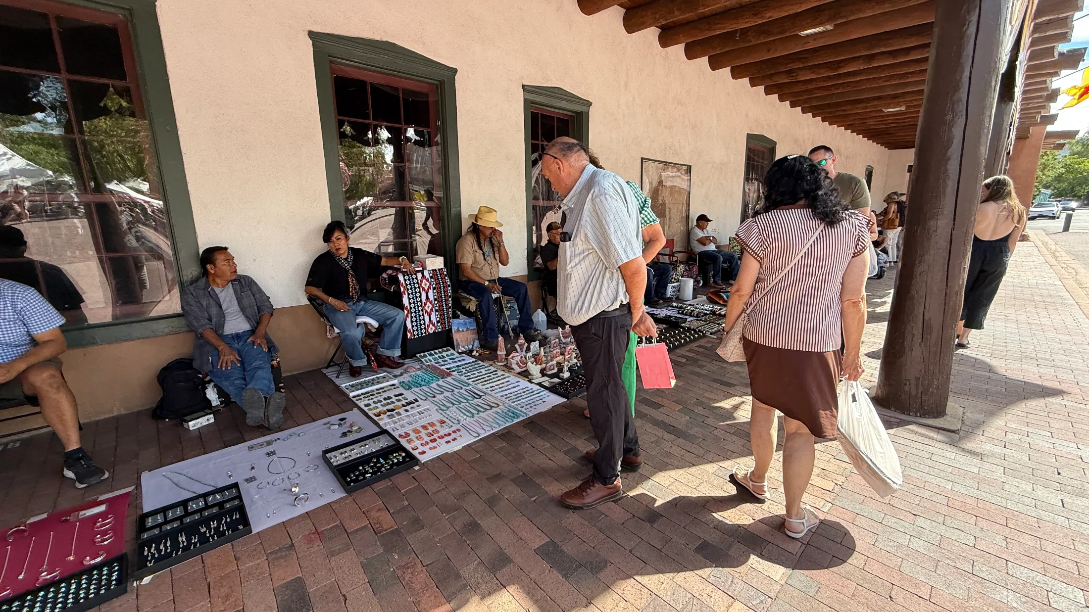 Native makers seated behind trays of silver, turquoise, coral, and beaded jewelry laid on blankets along the Palace of the Governors portal