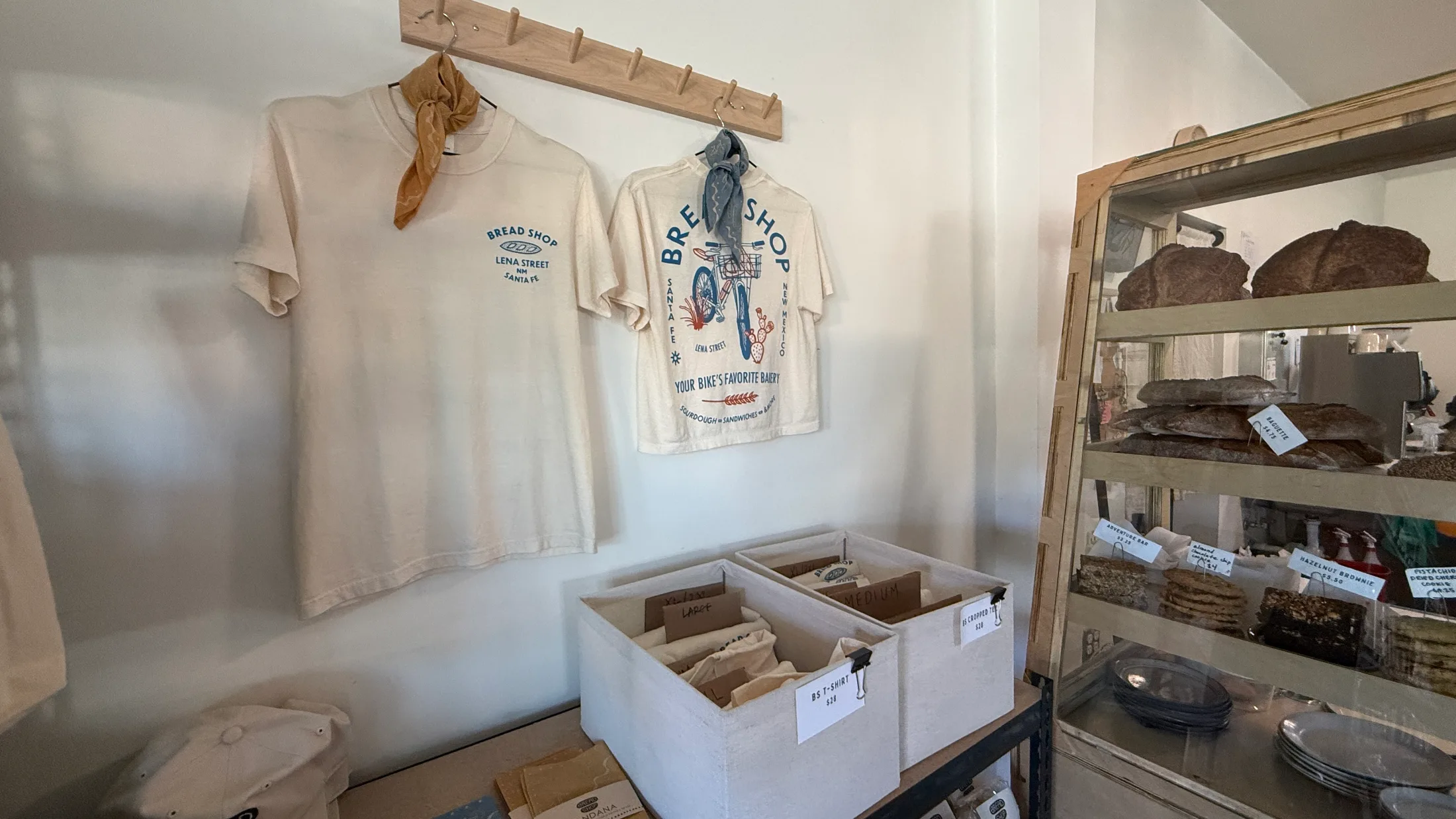 Bread Shop merchandise tees on a peg rack, including the Your Bike's Favorite Bakery shirt, with loaves on a shelf to the right