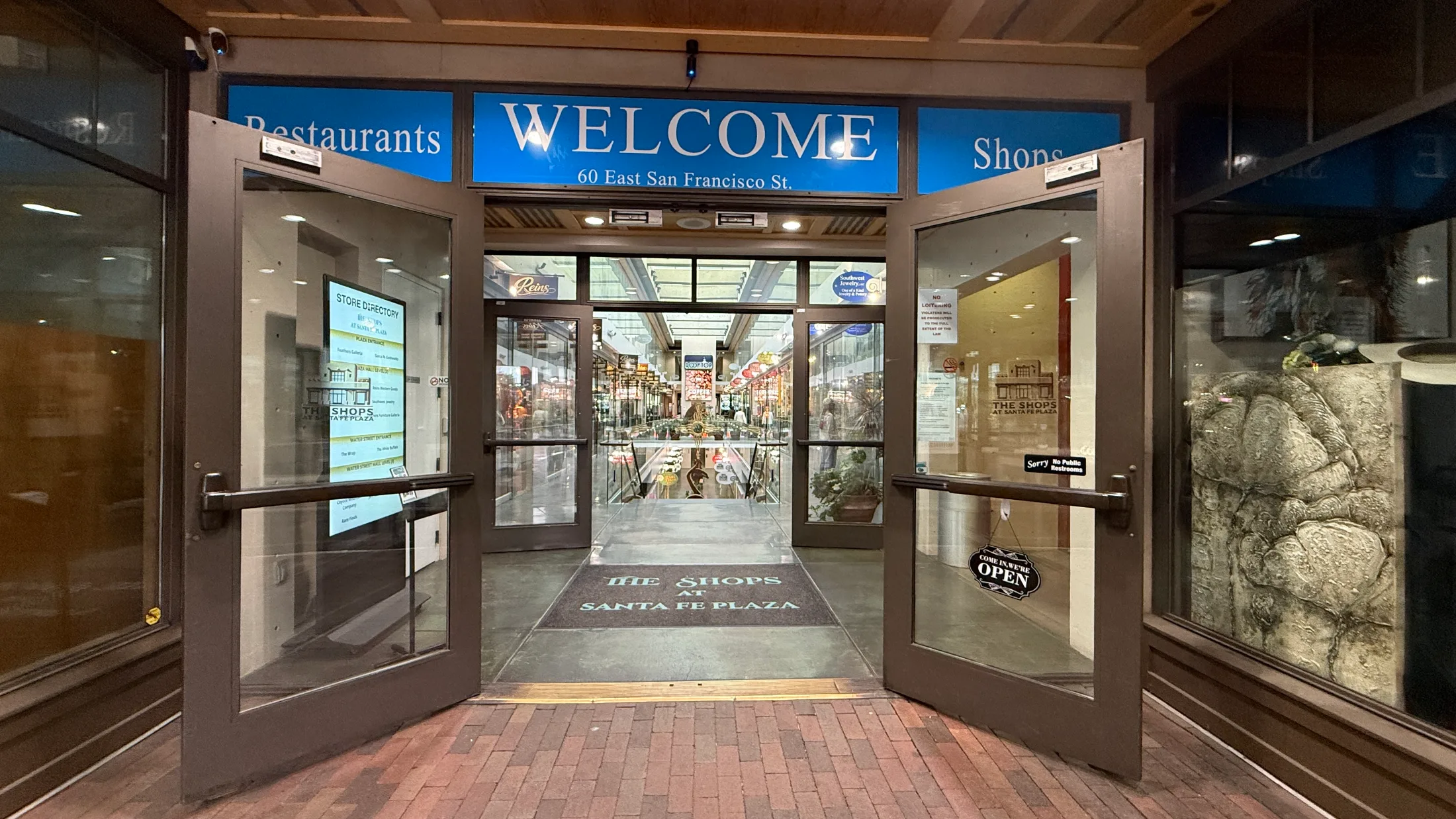 The entrance to the Shops at Santa Fe Plaza at 60 East San Francisco Street, with a blue WELCOME sign and a store directory visible through the glass doors
