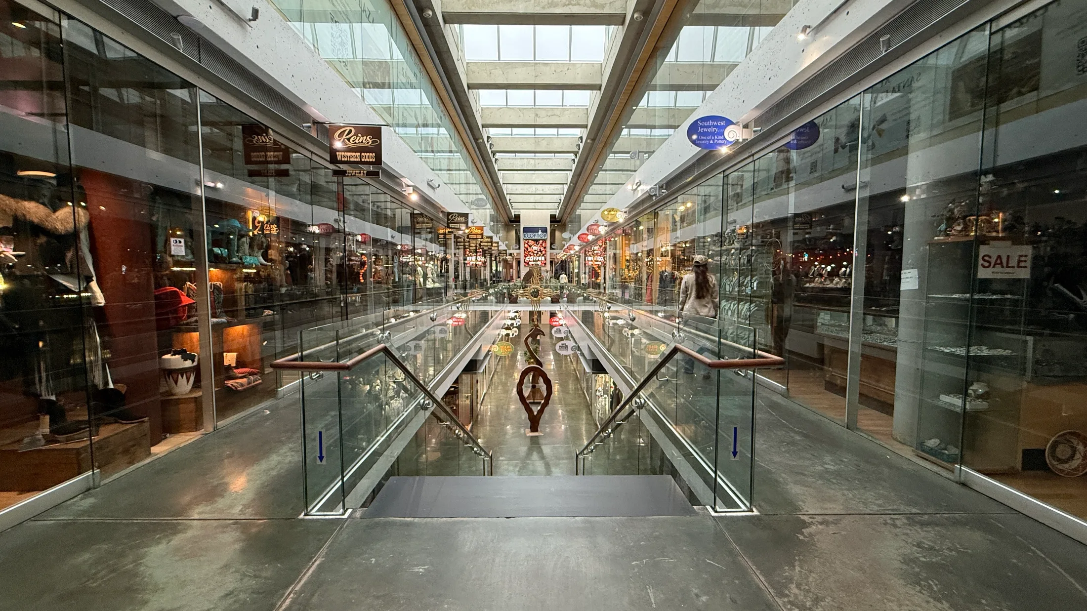 Looking down the center of the Shops at Santa Fe Plaza from the upper level staircase, with Reins Western Goods on the left, Southwest Jewelry on the right, and the full skylit arcade stretching ahead