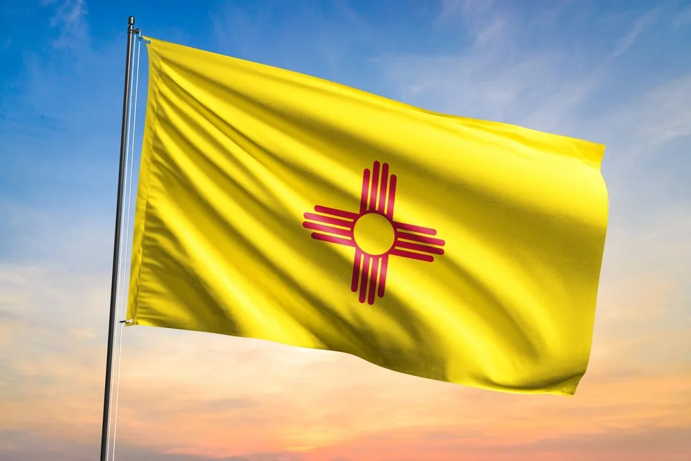 New Mexico state flag in yellow and red flying against a sunset sky, the Zia sun symbol prominent at center