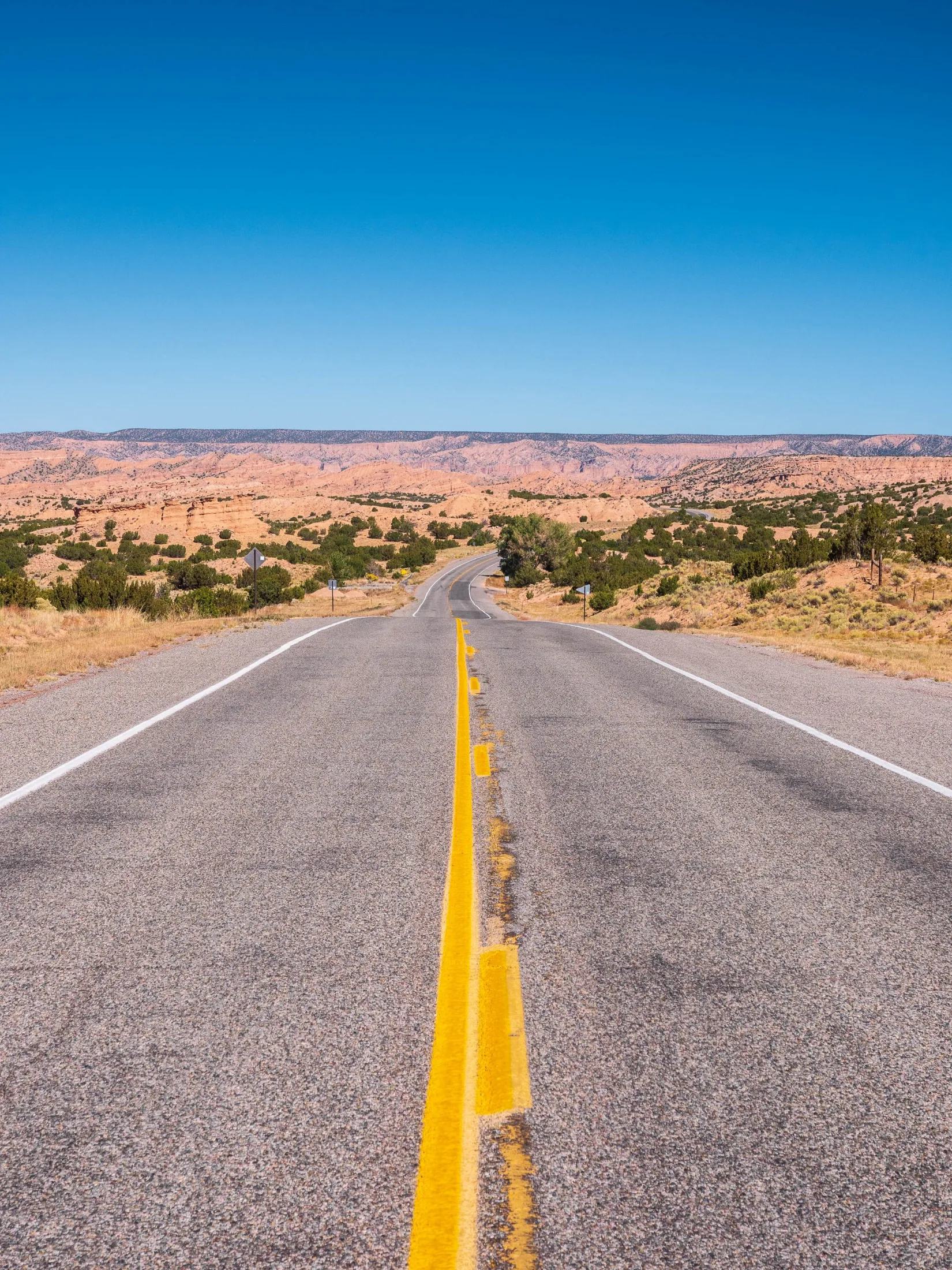Two-lane highway with a bold yellow center line stretching into the distance through red-rock desert and juniper scrub under a cobalt sky