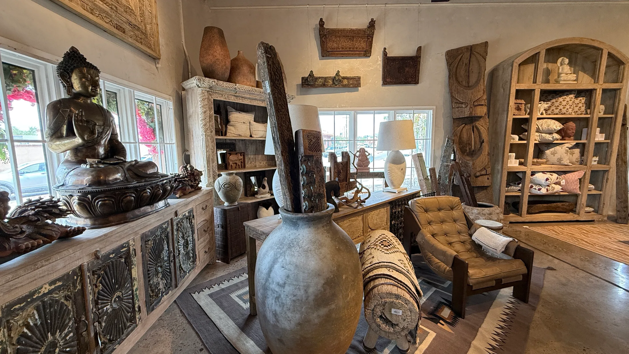 Detail vignette inside Sukhmani: a large bronze Buddha statue on a carved sideboard, a massive antique clay vessel, a tufted leather lounge chair, and weathered arched shelving filled with textiles and small sculptures