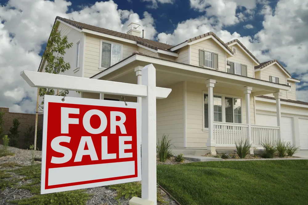 For sale sign in front of a large suburban home under a cloudy sky