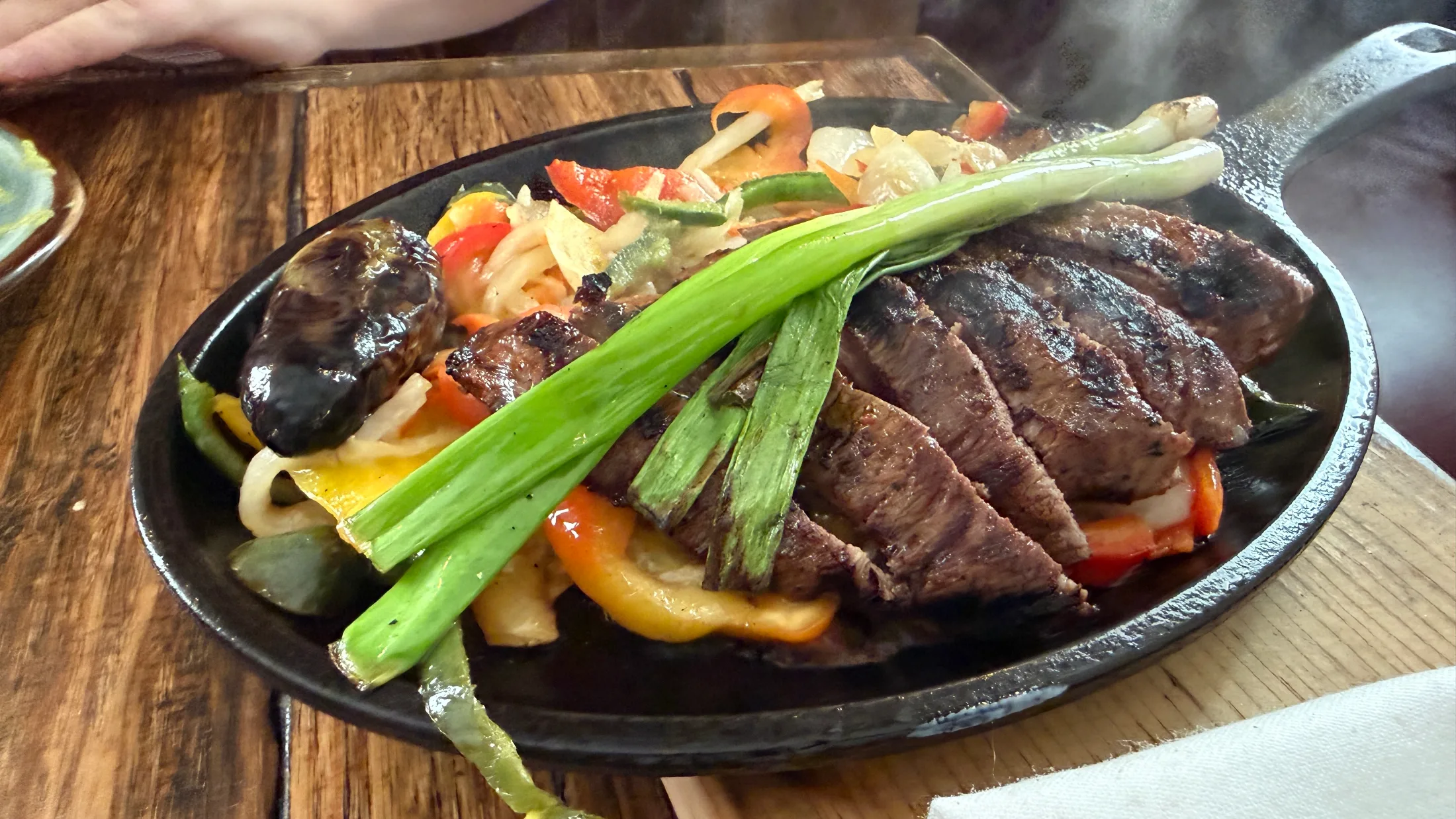Close-up of Paloma's sizzling steak fajita skillet with steam rising, charred vegetables and whole green onions