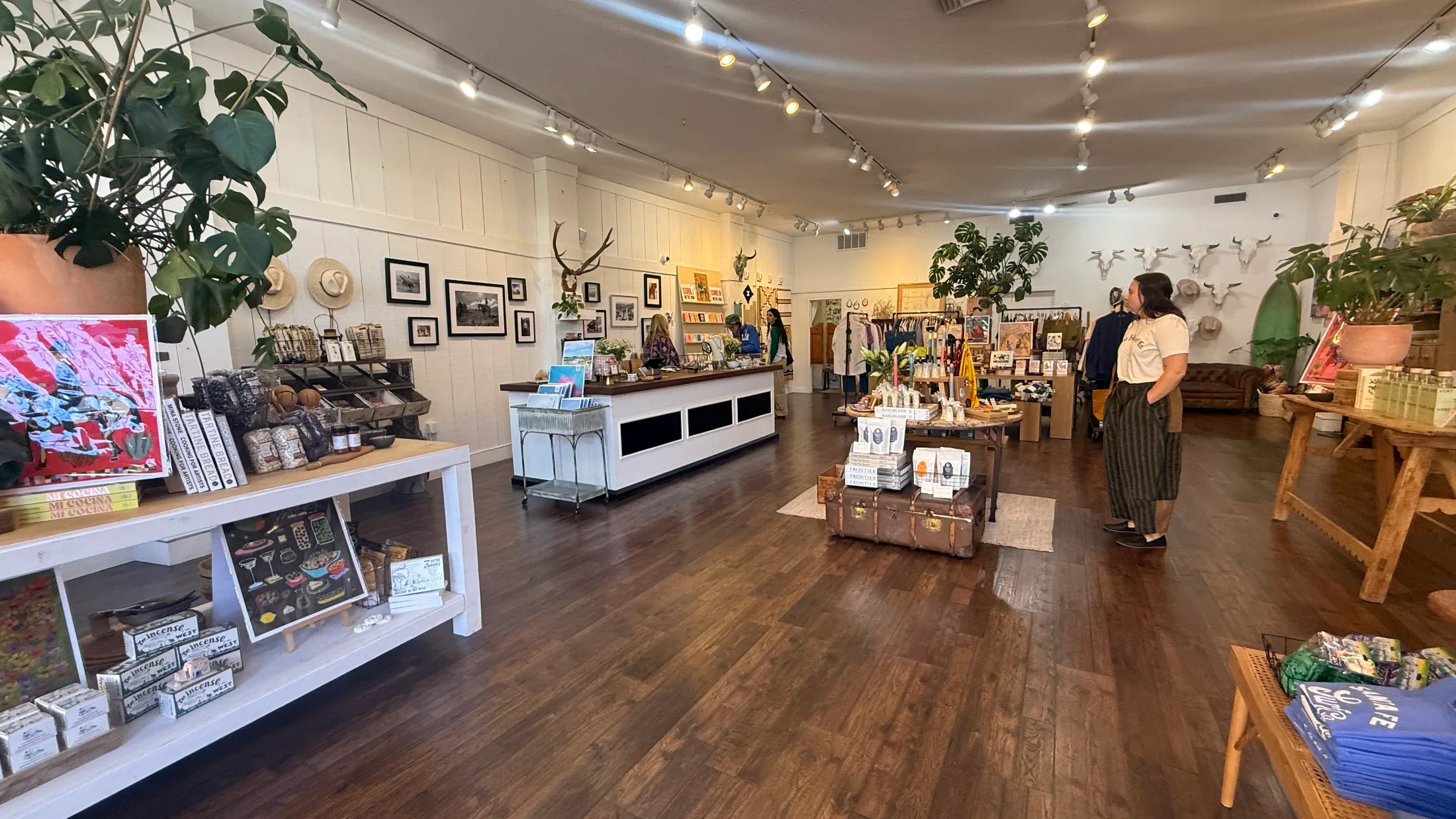 Interior overview of High Noon General Store showing the full room, white walls, dark wood floors, display tables, clothing rack, and shoppers browsing