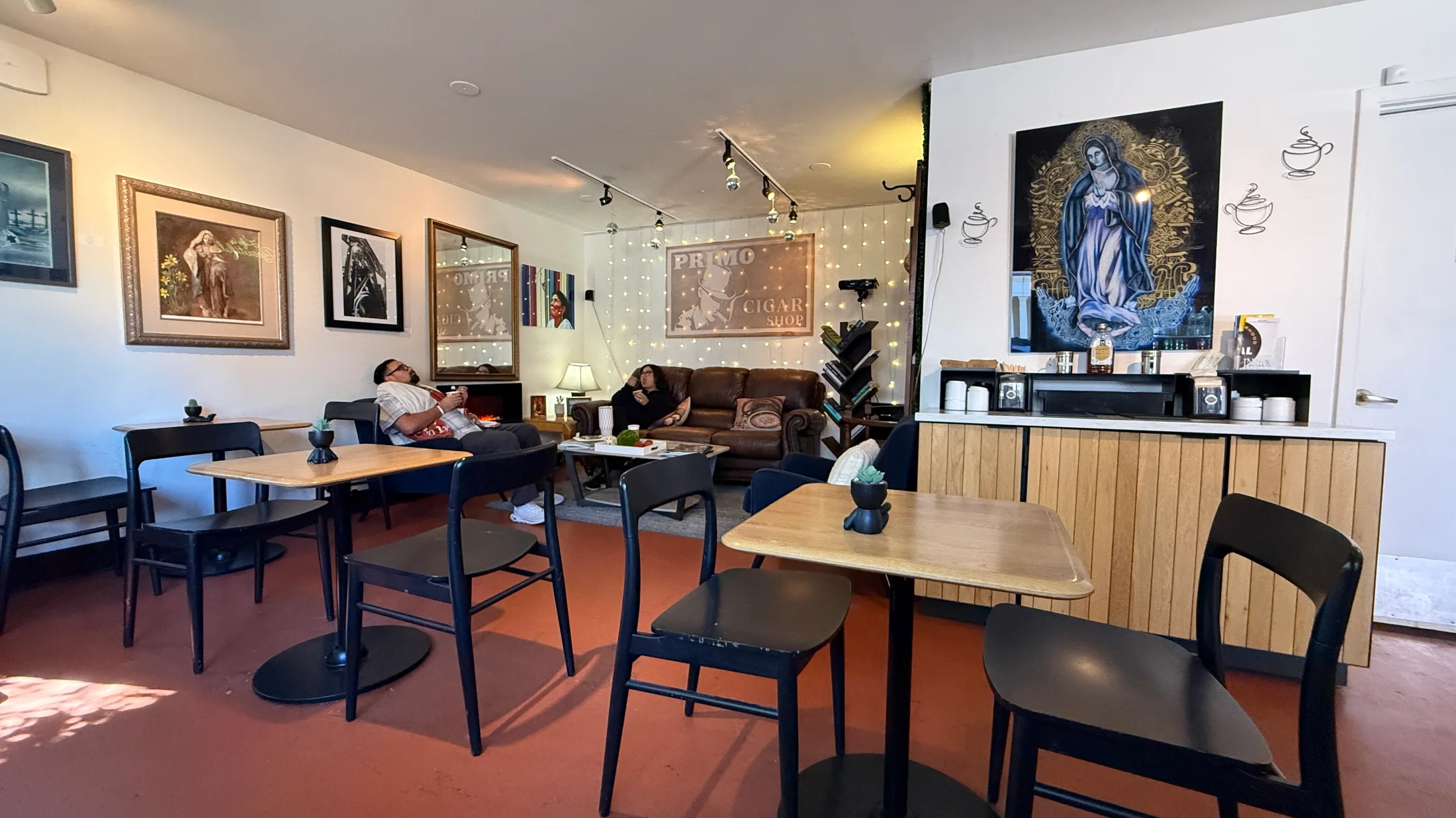 Lounge area at Santa Fe Biscochito Company with a leather couch under the original Primo Cigar Shop sign, string lights, a devotional painting of Our Lady, and cafe tables in the foreground
