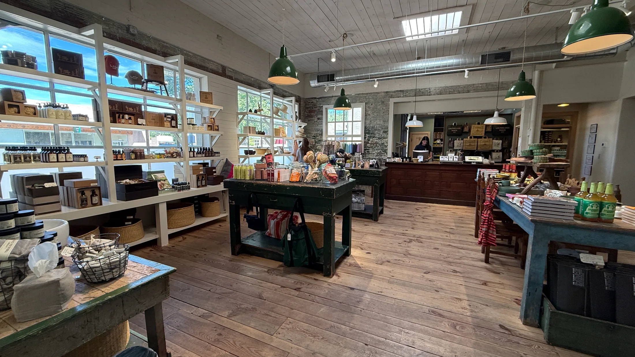 Interior of Farm Shop Norte showing white shelving units, green farm tables loaded with pantry goods, candles, and body care products under green pendant lights