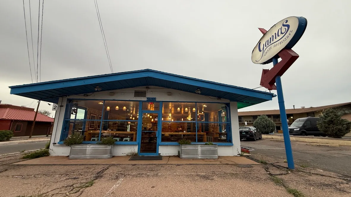 Yamas Greek Rotisserie exterior at dusk, address 2411 visible on the blue-trimmed facade, warm light glowing through the front windows