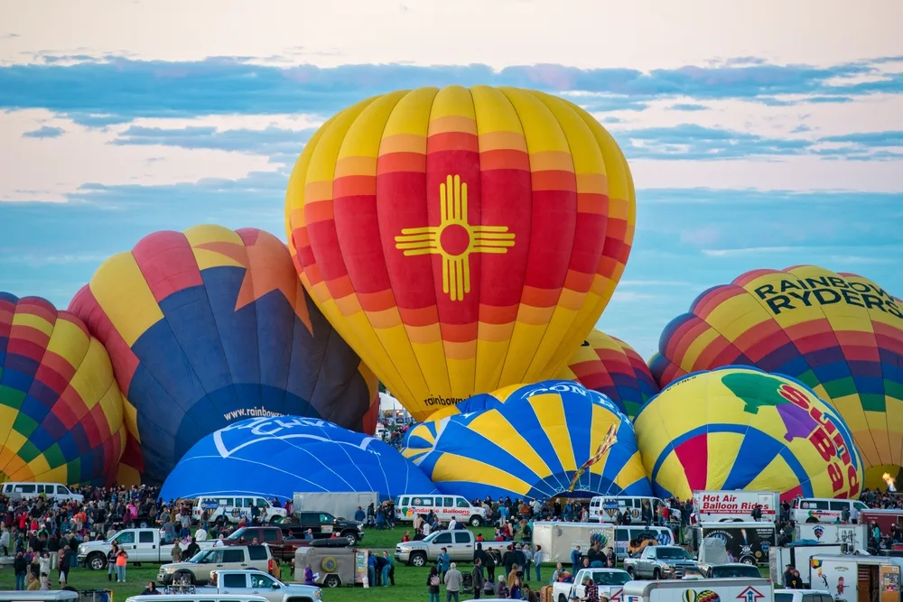 Dozens of hot air balloons inflating on a field at dawn, one bearing the Zia symbol in gold on deep red and yellow panels, crews and crowds surrounding them