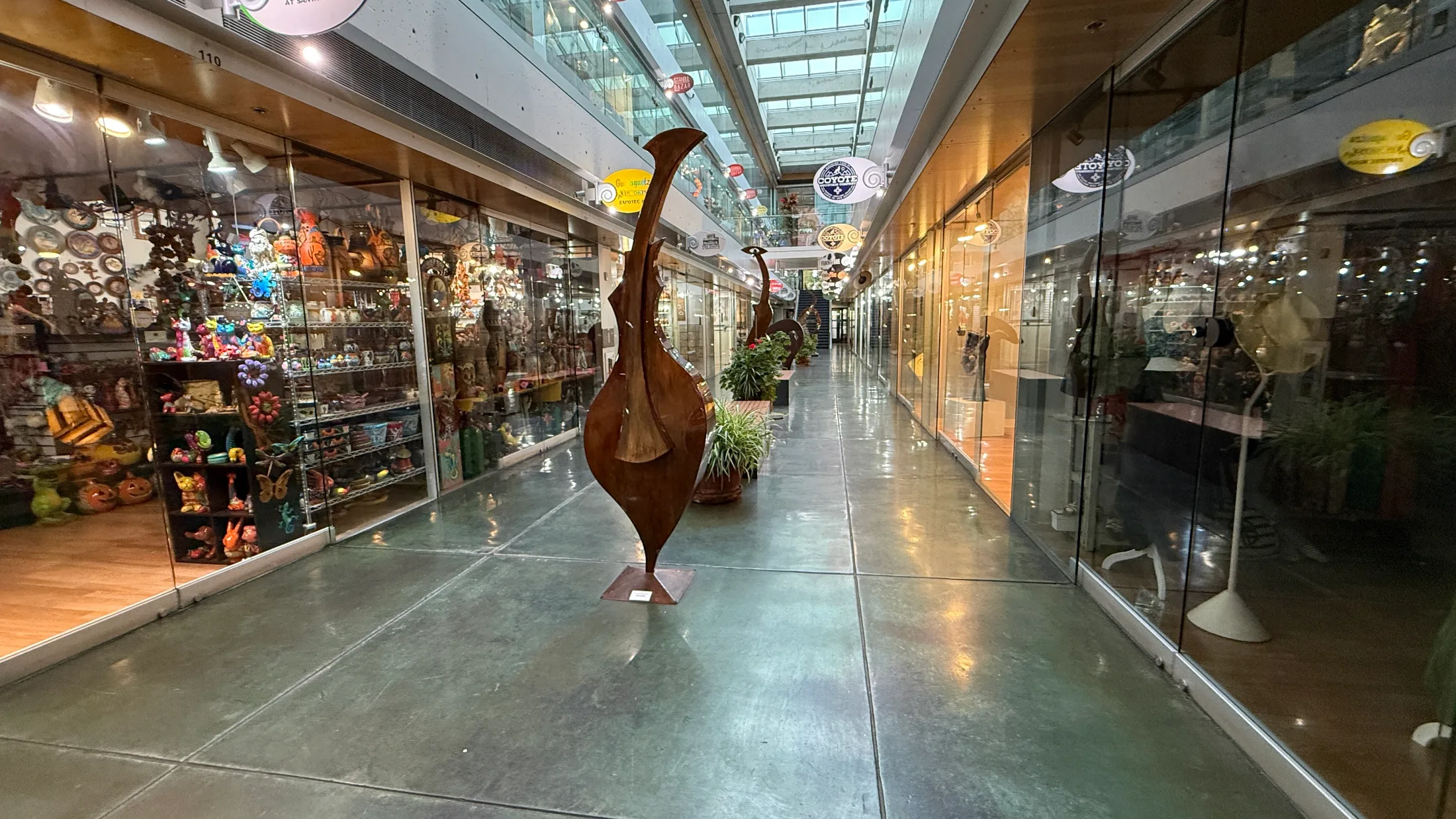 The lower-level corridor of the Shops at Santa Fe Plaza with rust-patina metal sculptures standing between storefronts, a skylit ceiling above, and a colorful folk-art shop visible on the left