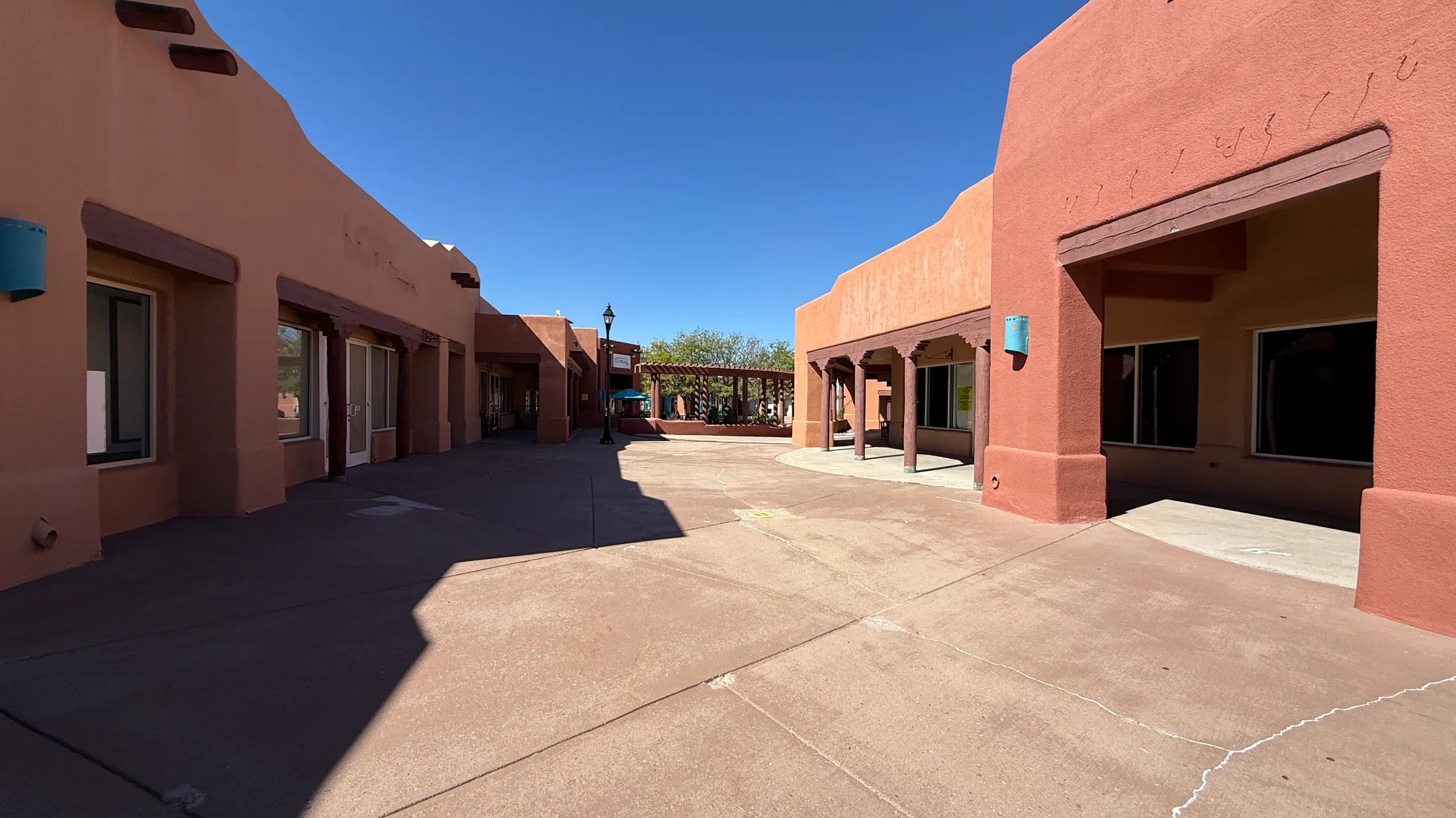Empty courtyard walkway flanked by adobe-style storefronts, with a pergola visible in the background and several vacant units