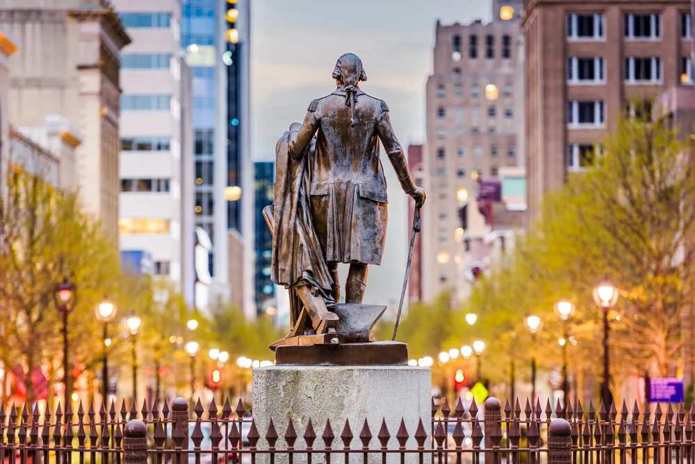 Bronze statue of a founding-era figure looking down a tree-lined Raleigh boulevard at dusk, city buildings glowing behind