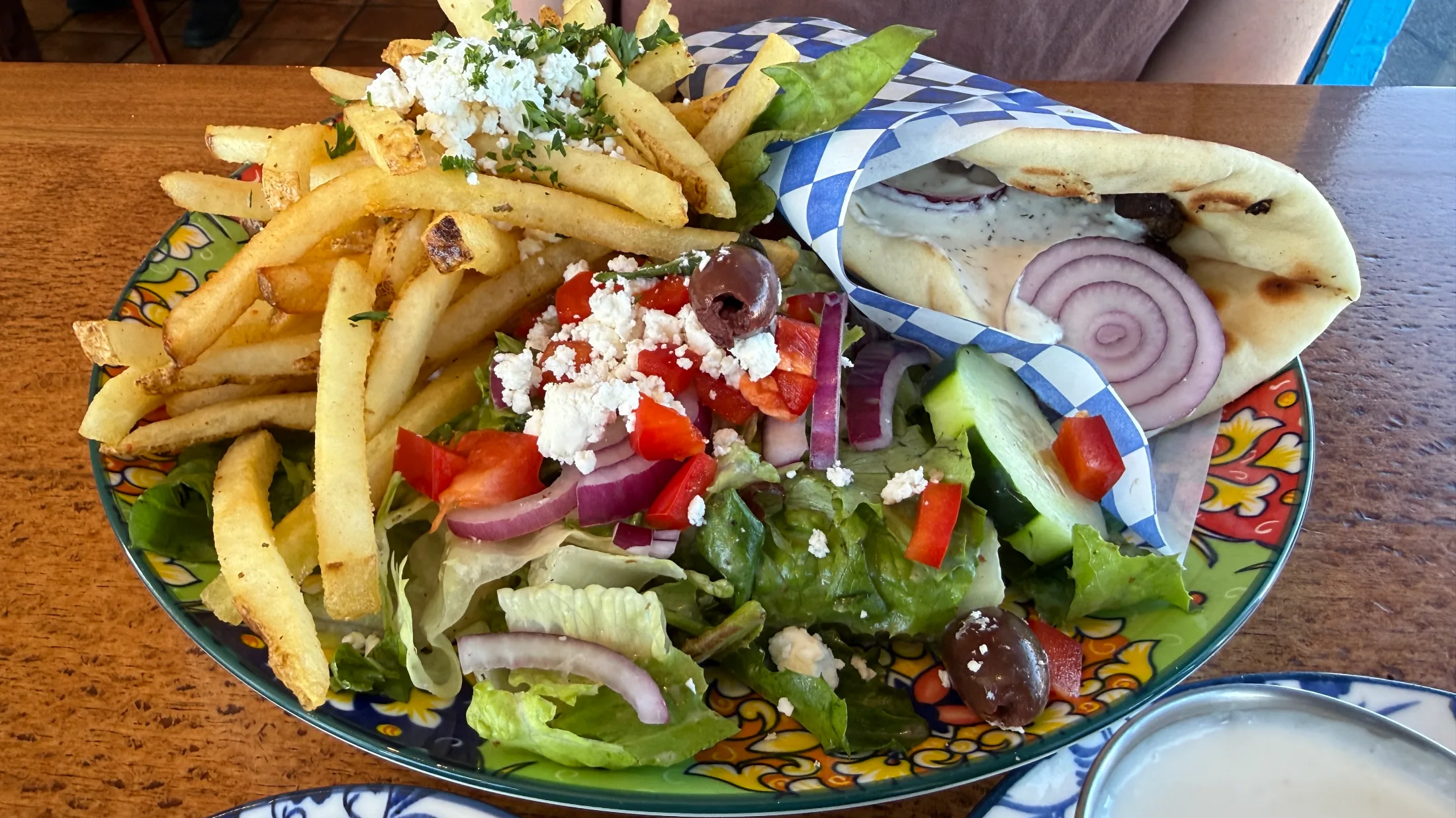 A gyro wrapped in blue-and-white checkered paper alongside Greek salad with feta and kalamata olives, and a pile of fries dusted with crumbled feta