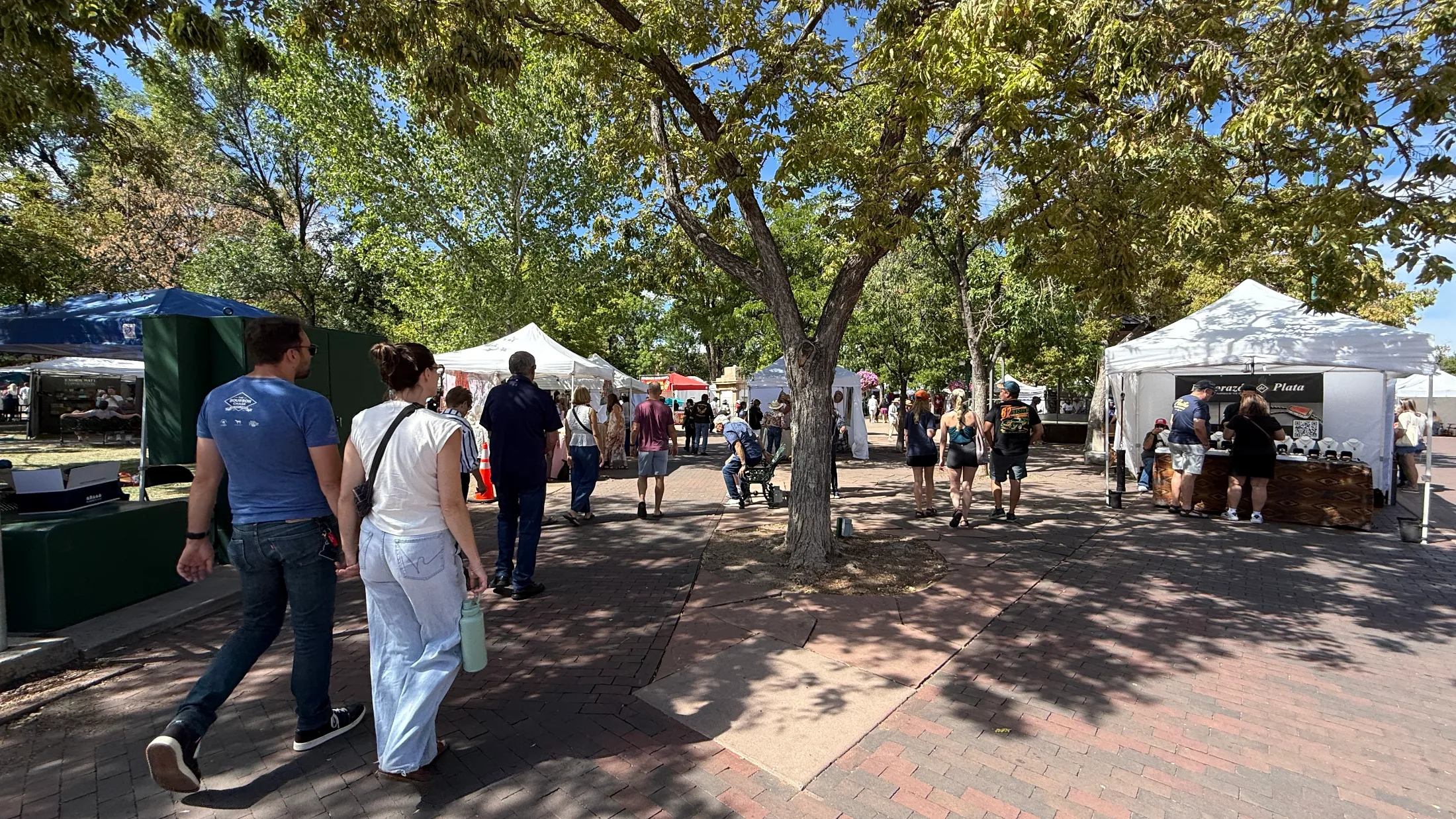 Couples and families walk through artisan vendor tents filling the shaded brick paths of the Santa Fe Plaza park on a weekend morning