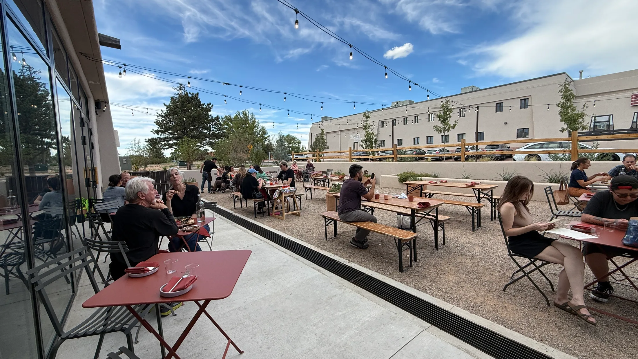 Tender Fire patio full of diners at communal bench tables and red bistro tables under string lights, glass wall of the restaurant to the left, large stucco building visible across the lot