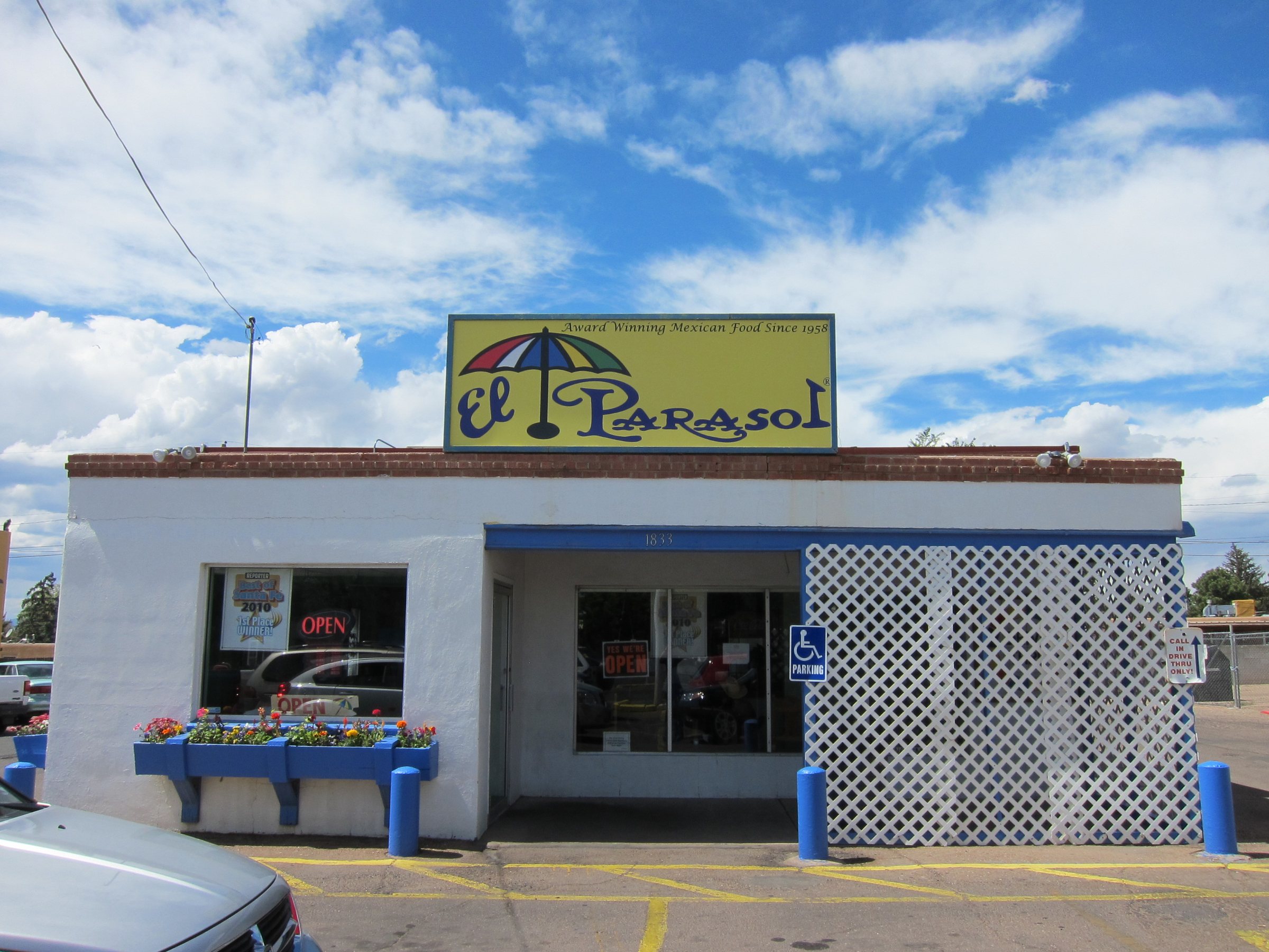A white El Parasol building with a yellow sign showing a striped umbrella, the number 1833 over the door and blue flower boxes under the window