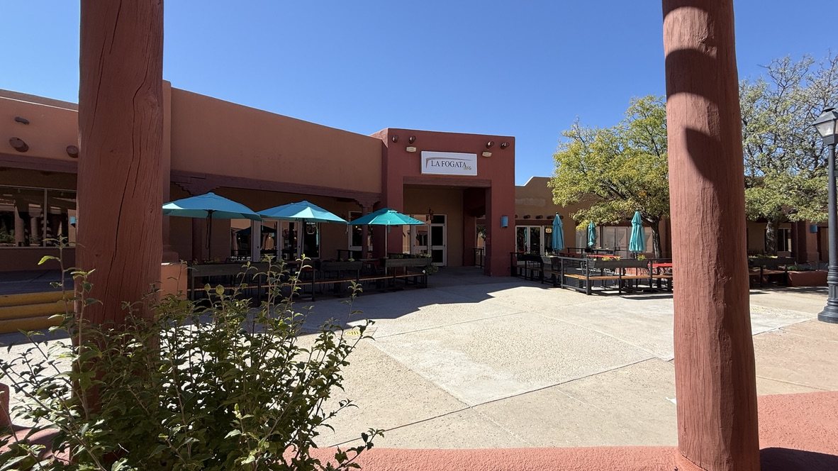 A courtyard with tan stucco buildings, wooden posts and teal umbrellas under a blue sky