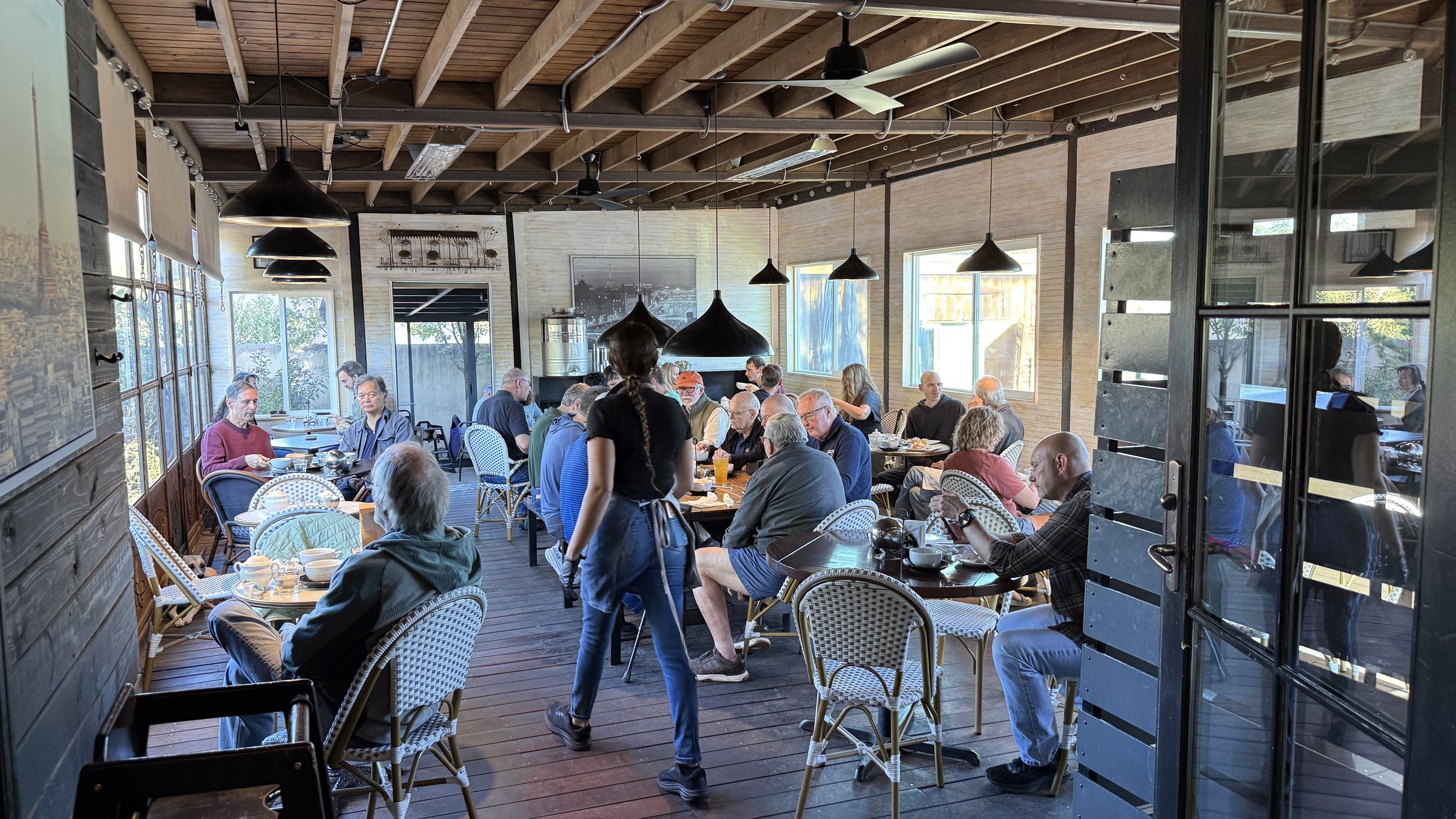 A busy covered patio with a wood beam ceiling, black pendant lamps and woven bistro chairs at round tables