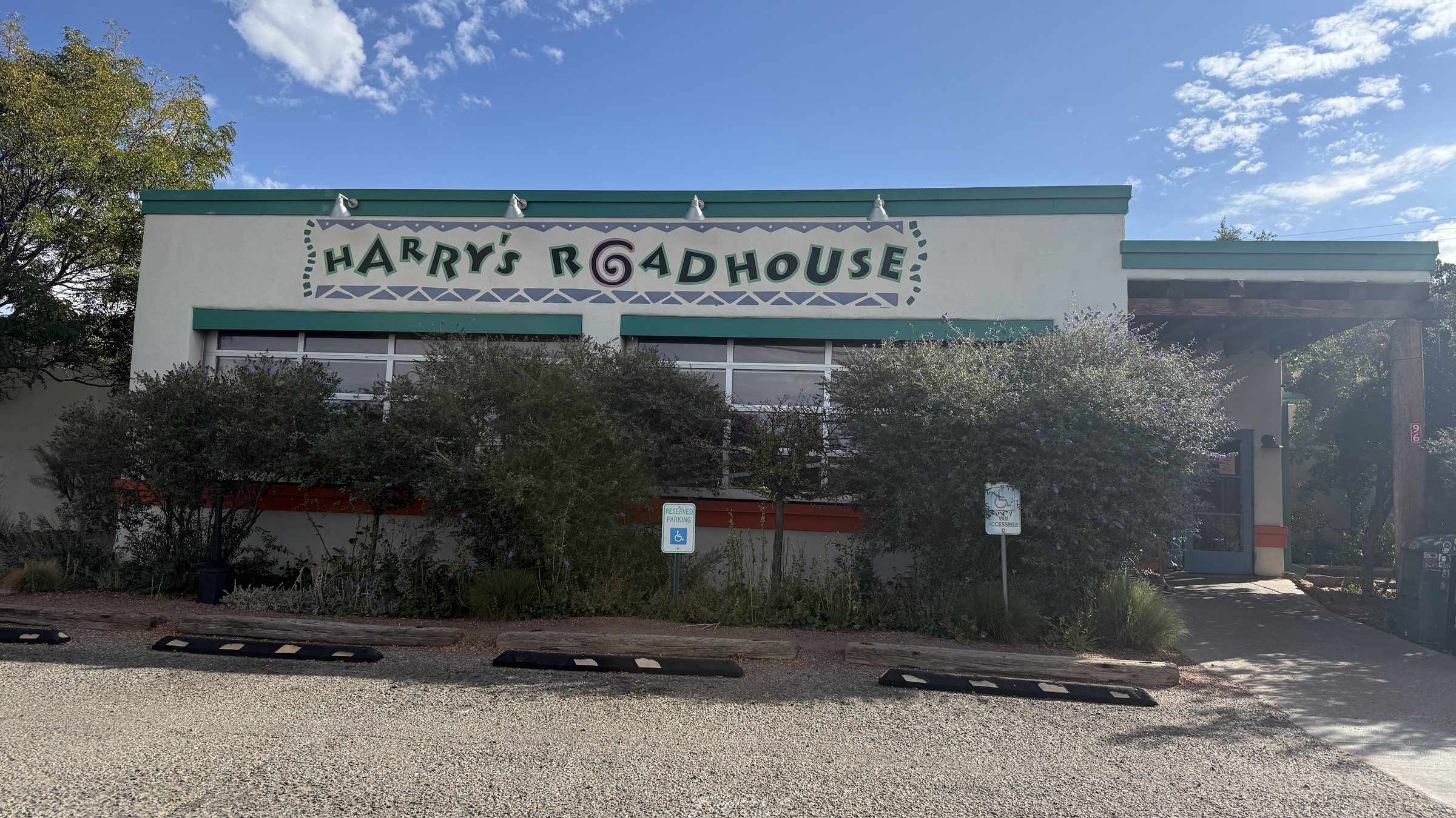 The white Harry's Roadhouse building with green trim and its sign, behind shrubs and a gravel lot under a blue sky