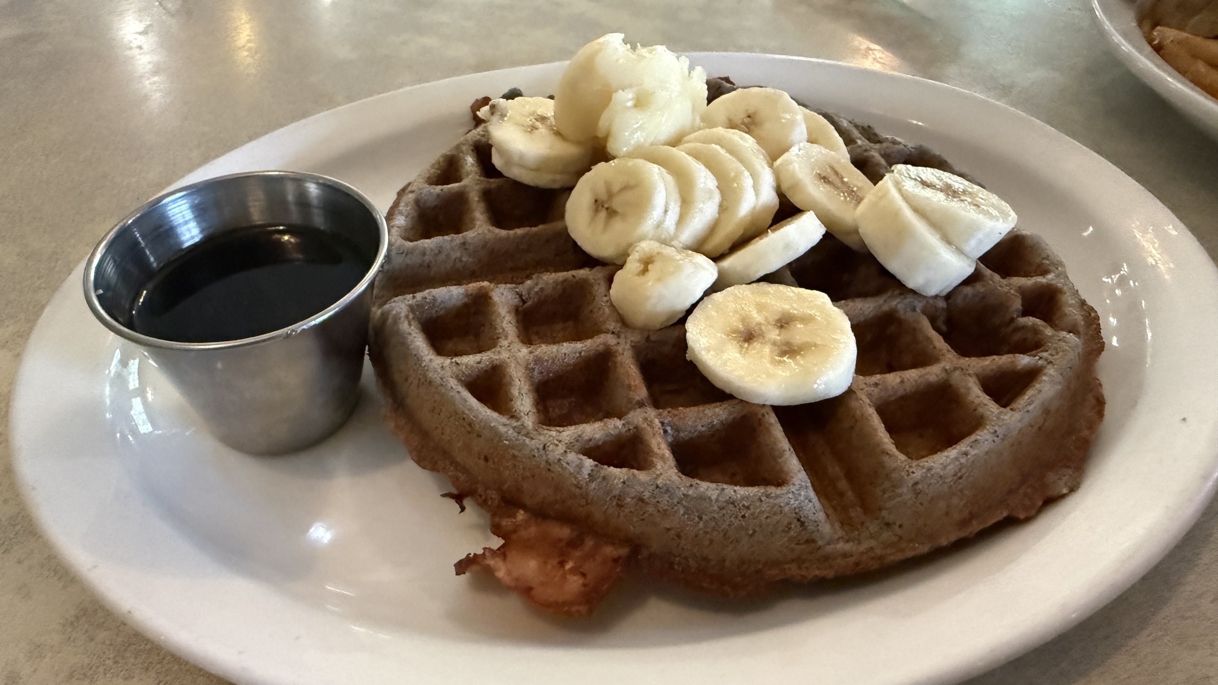 A dark waffle topped with sliced bananas beside a metal cup of syrup