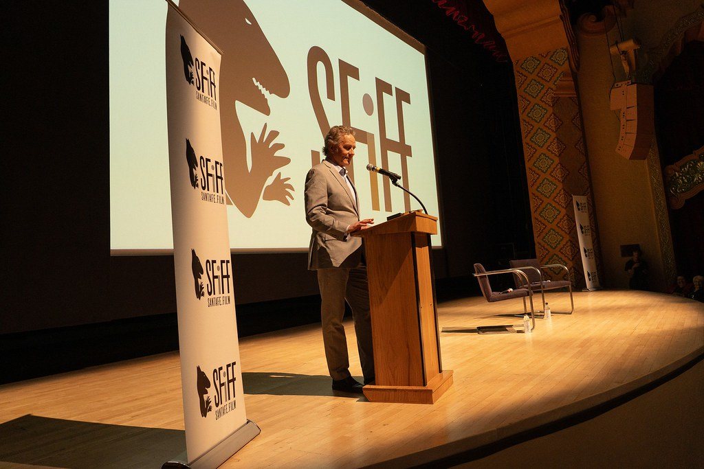 Man in a suit at a wooden podium on a theater stage with a logo on the screen behind him