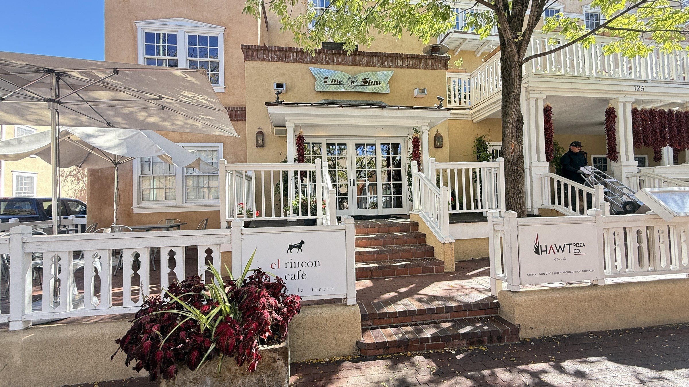 The front of Hotel Chimayó with a white wooden railing, steps up to a porch, and signs for El Rincón Café and HAWT on the railing, with a tree and a blue sky