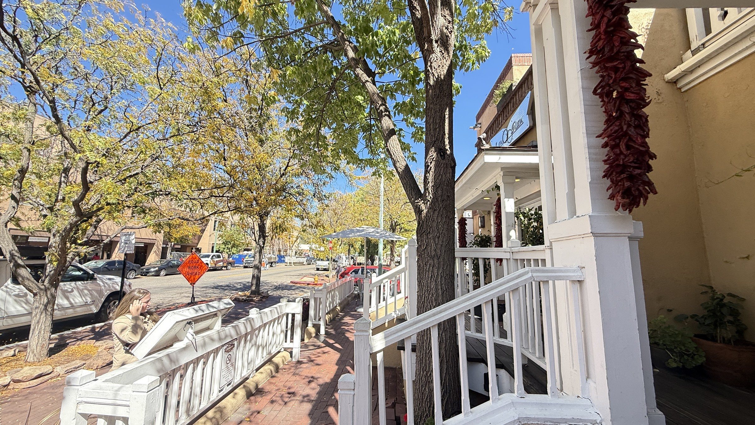 A brick sidewalk along Washington Avenue with white railings, a hanging red chile ristra and trees with yellow leaves