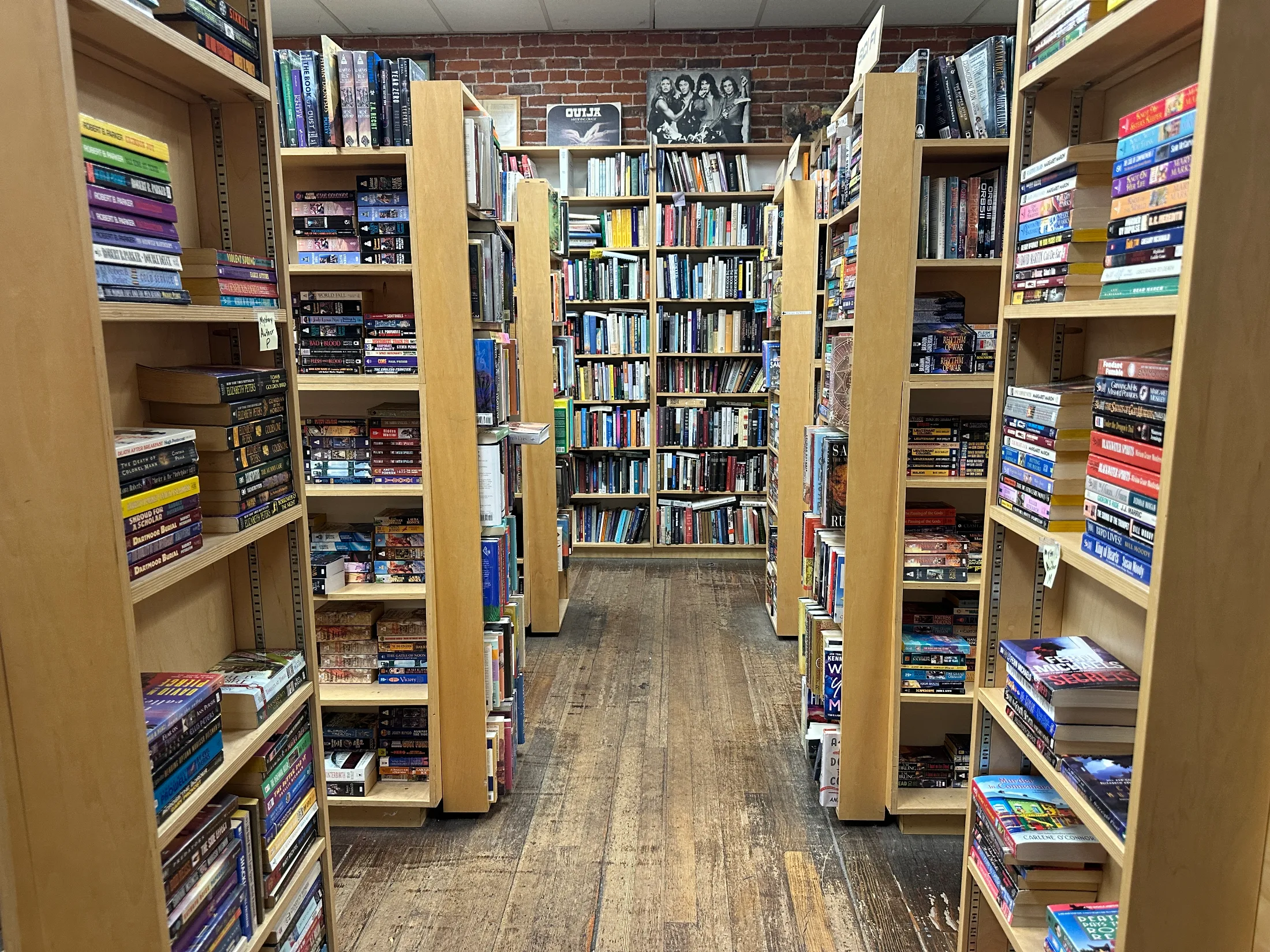 Dense shelving in a back room of The Bookstore packed with paperback fiction and genre titles, with a Ouija board visible on top of a shelf