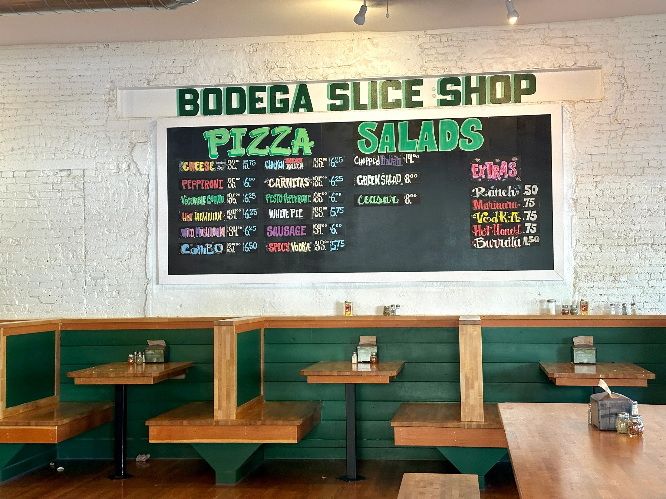 Bodega Slice Shop chalkboard menu showing pizza, salad, and extras prices on a white-painted brick wall above green booths