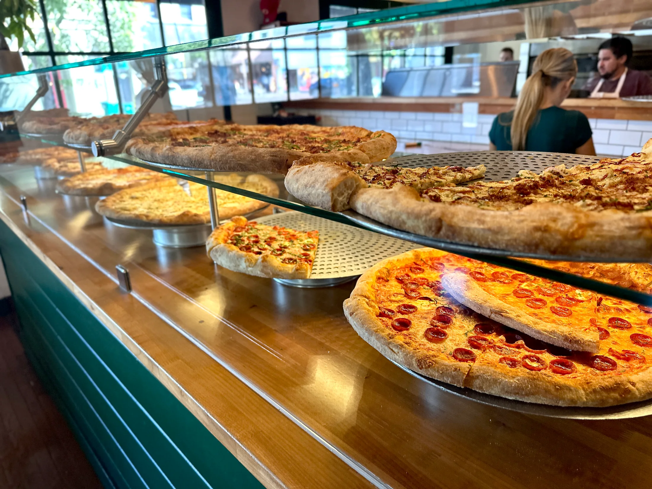 Long counter display of giant New York-style pizza pies at Bodega Slice Shop, with staff visible in the background